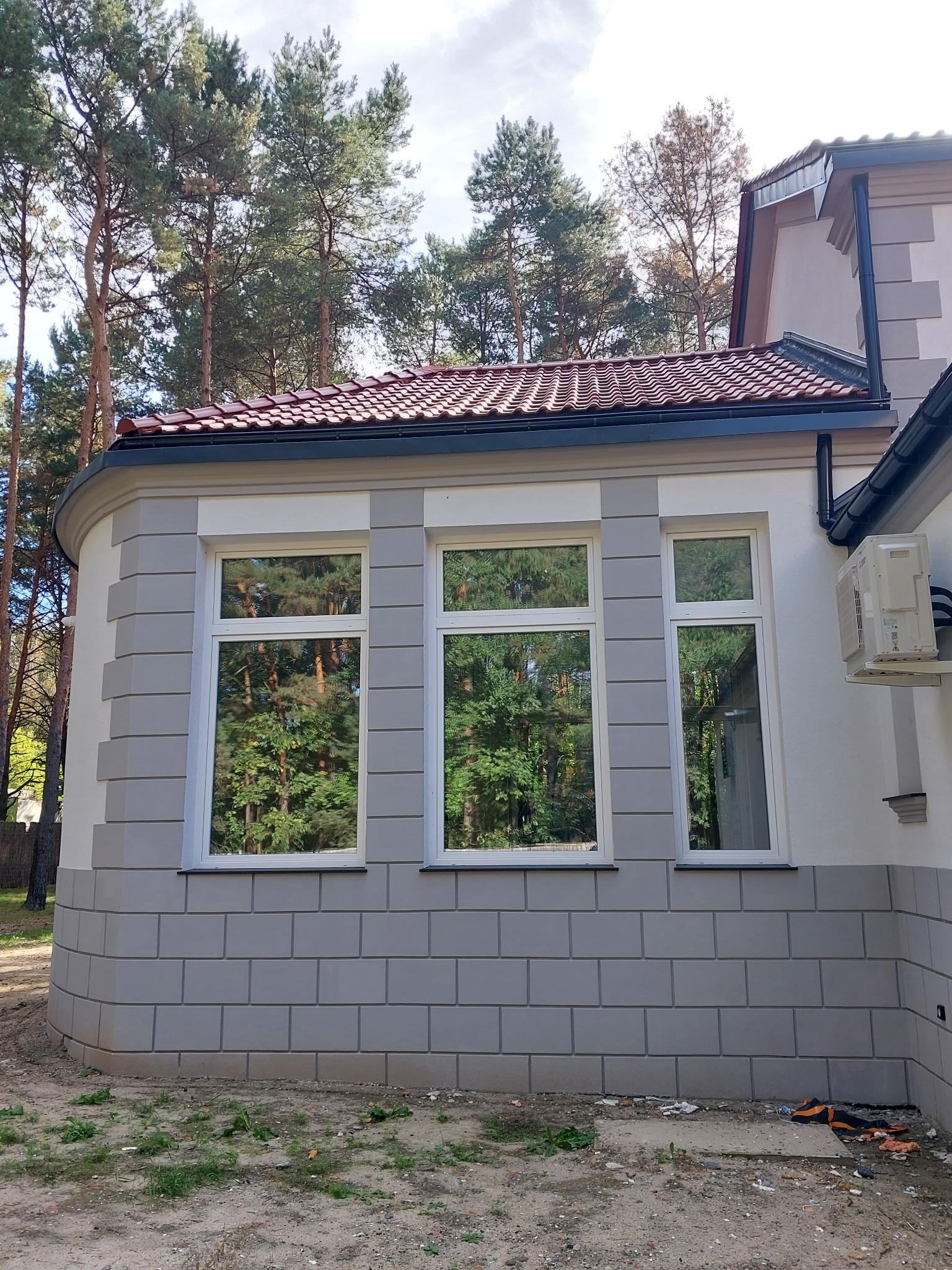 Elewacja budynku z trzema oknami i dekoracyjnym tynkiem imitującym cegłę, z widoczną jednostką zewnętrzną klimatyzacji i odbiciem drzew w szybach okiennych.