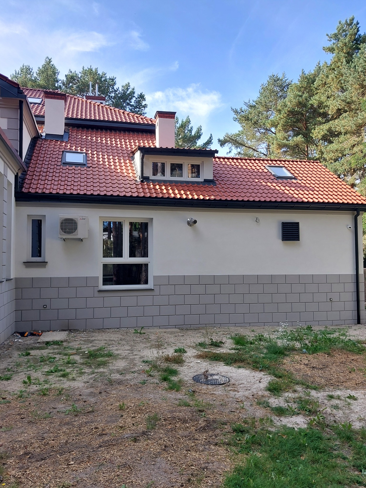 Wykończona elewacja domu jednorodzinnego z czerwoną dachówką, szarą podmurówką imitującą cegłę, klimatyzacją i wentylacją, widok z zewnątrz w słoneczny dzień.
