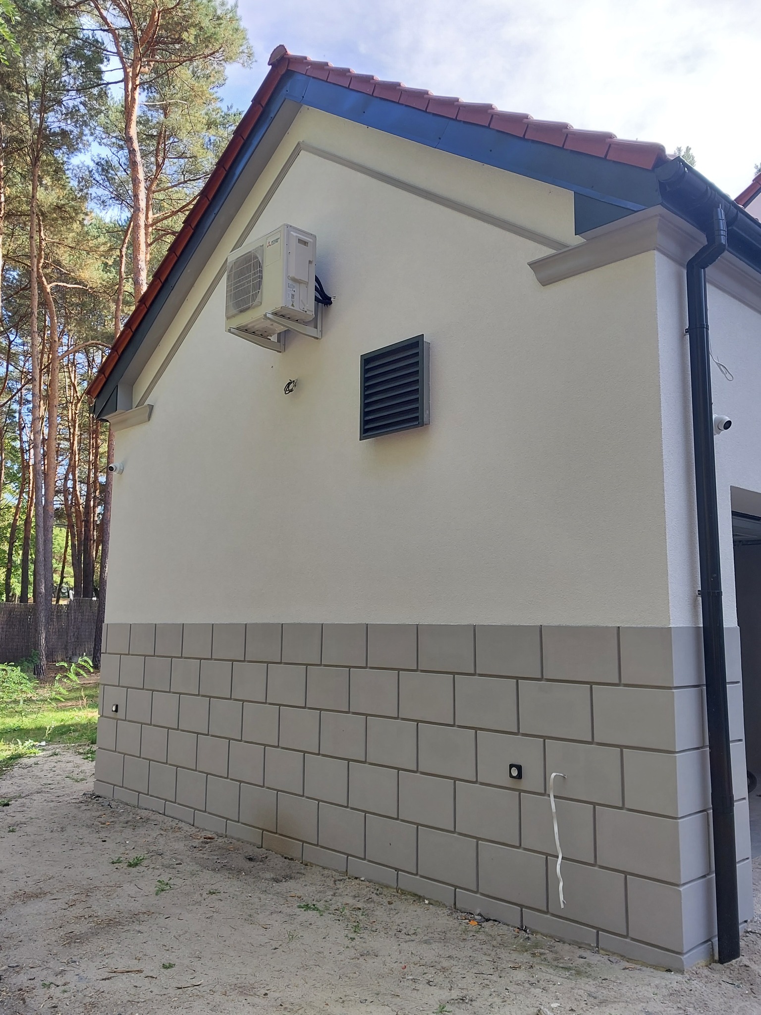 Ściana domu z nową elewacją, widoczny klimatyzator, kratka wentylacyjna i kamera monitoringu. Część elewacji imituje mur z szarych cegieł.