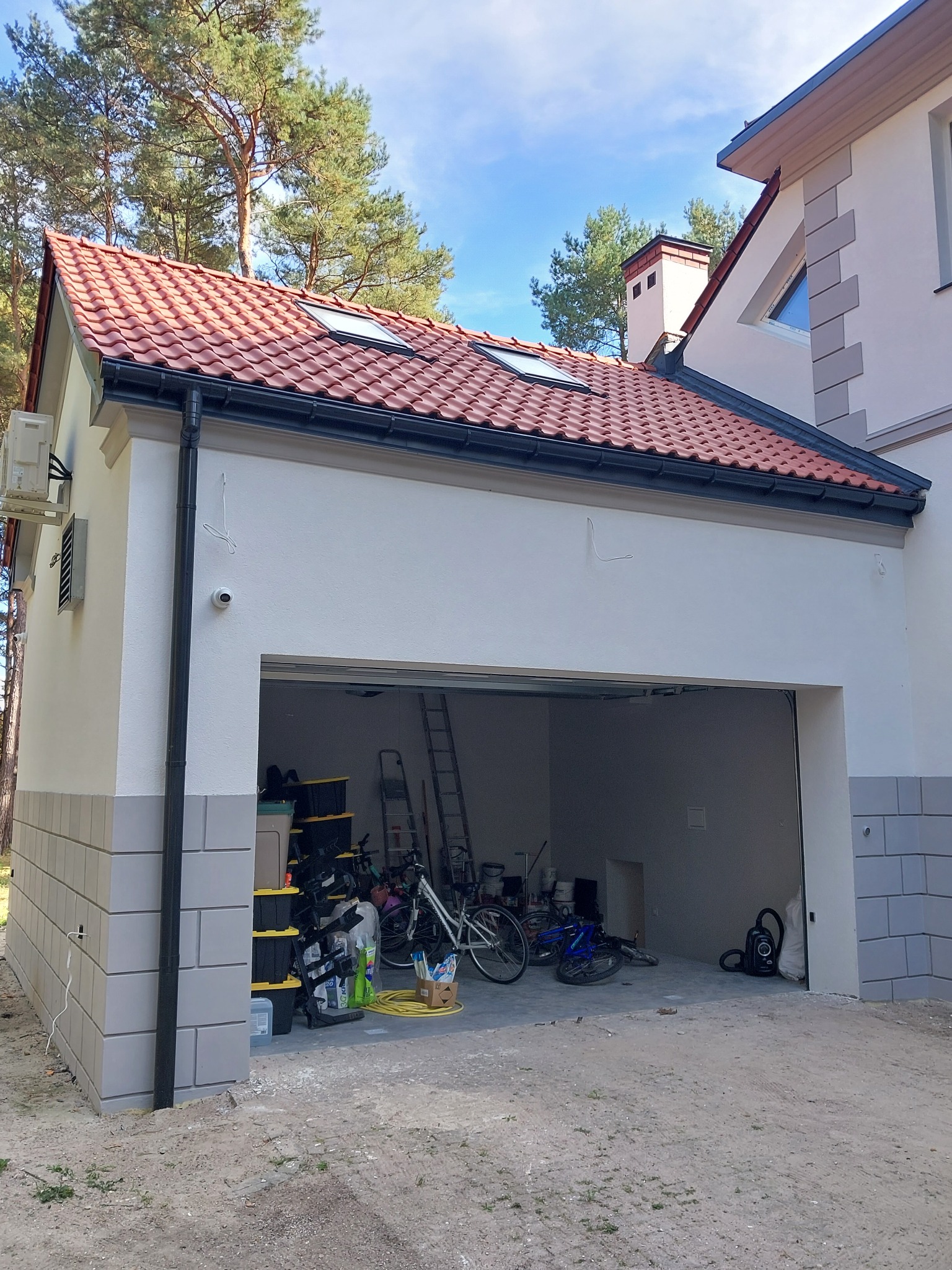 Nowo wybudowany garaż z czerwoną dachówką i jasną elewacją, widoczny bałagan wewnątrz, drabina i rowery.