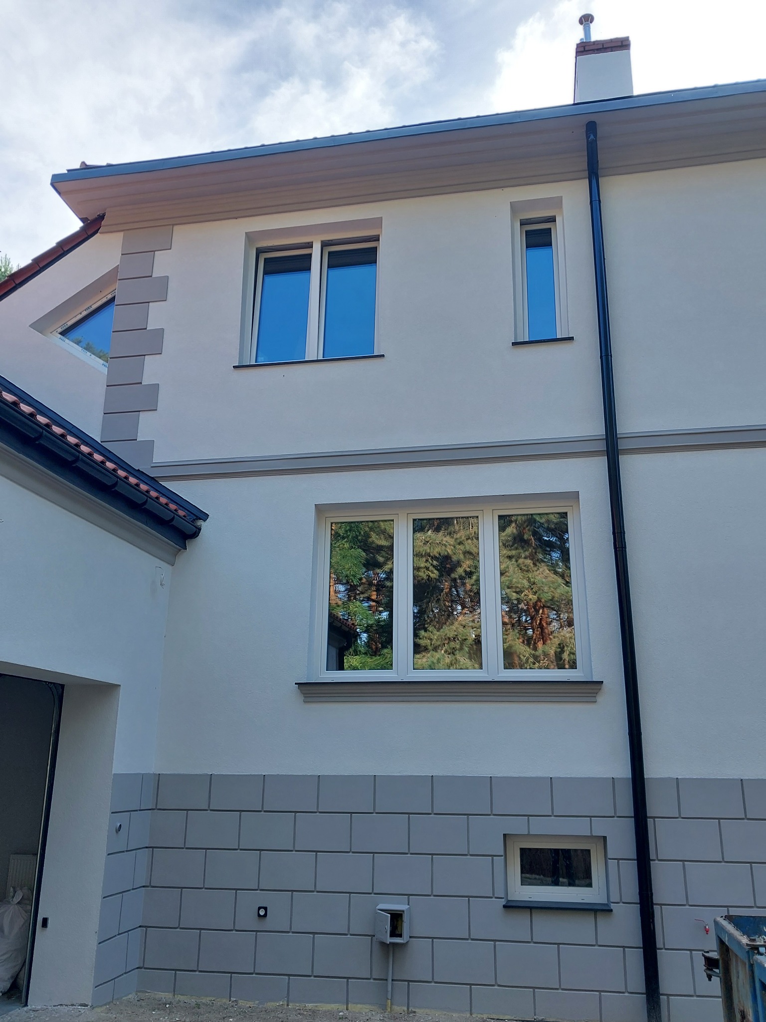 Odnowiona elewacja domu jednorodzinnego z szarym cokołem imitującym cegłę, białymi ścianami i nowymi oknami, widok z zewnątrz w słoneczny dzień.