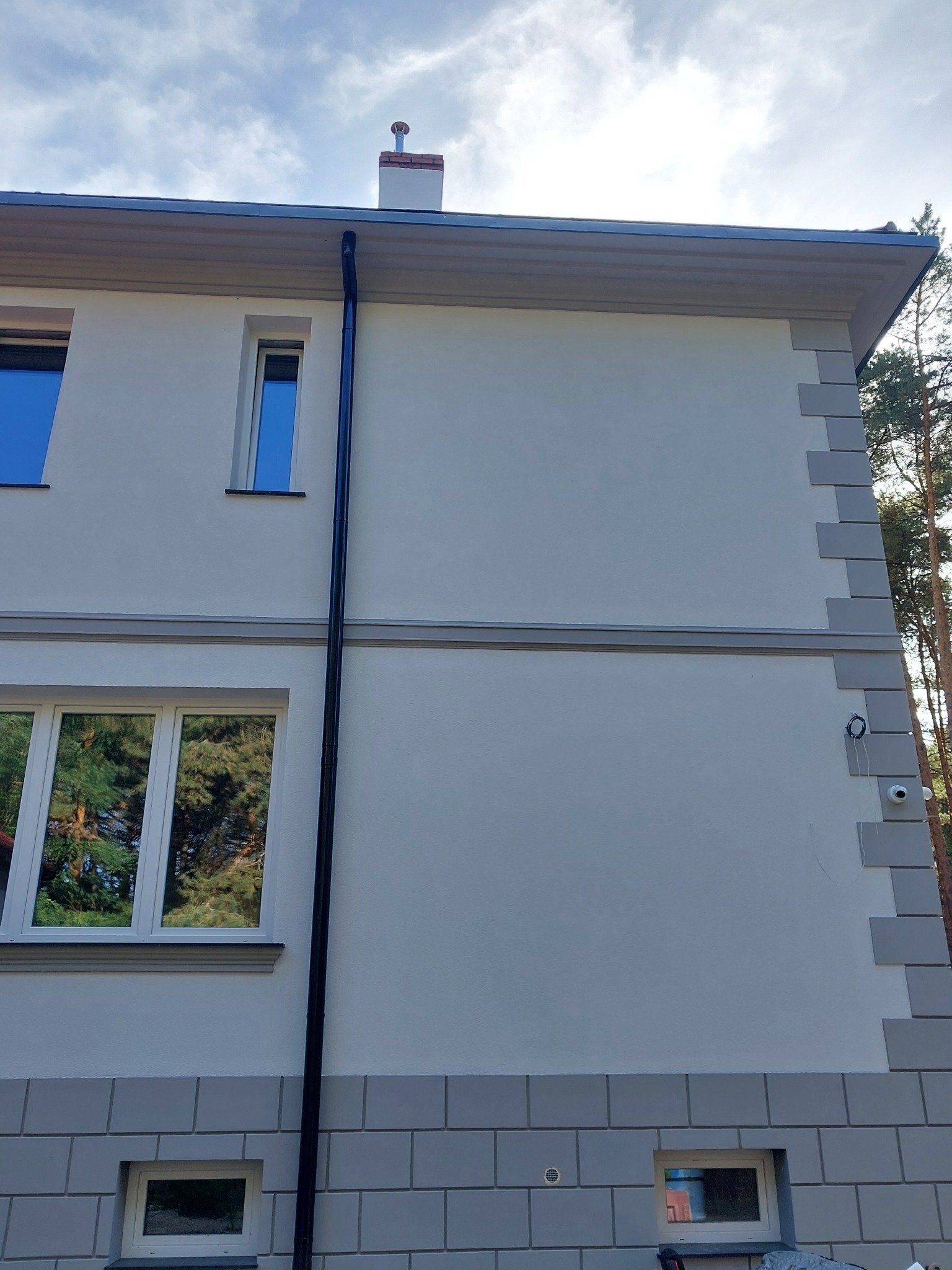 Dwupiętrowy dom z jasną elewacją, szarymi detalami architektonicznymi i czarną rynną, widok z zewnątrz w słoneczny dzień.