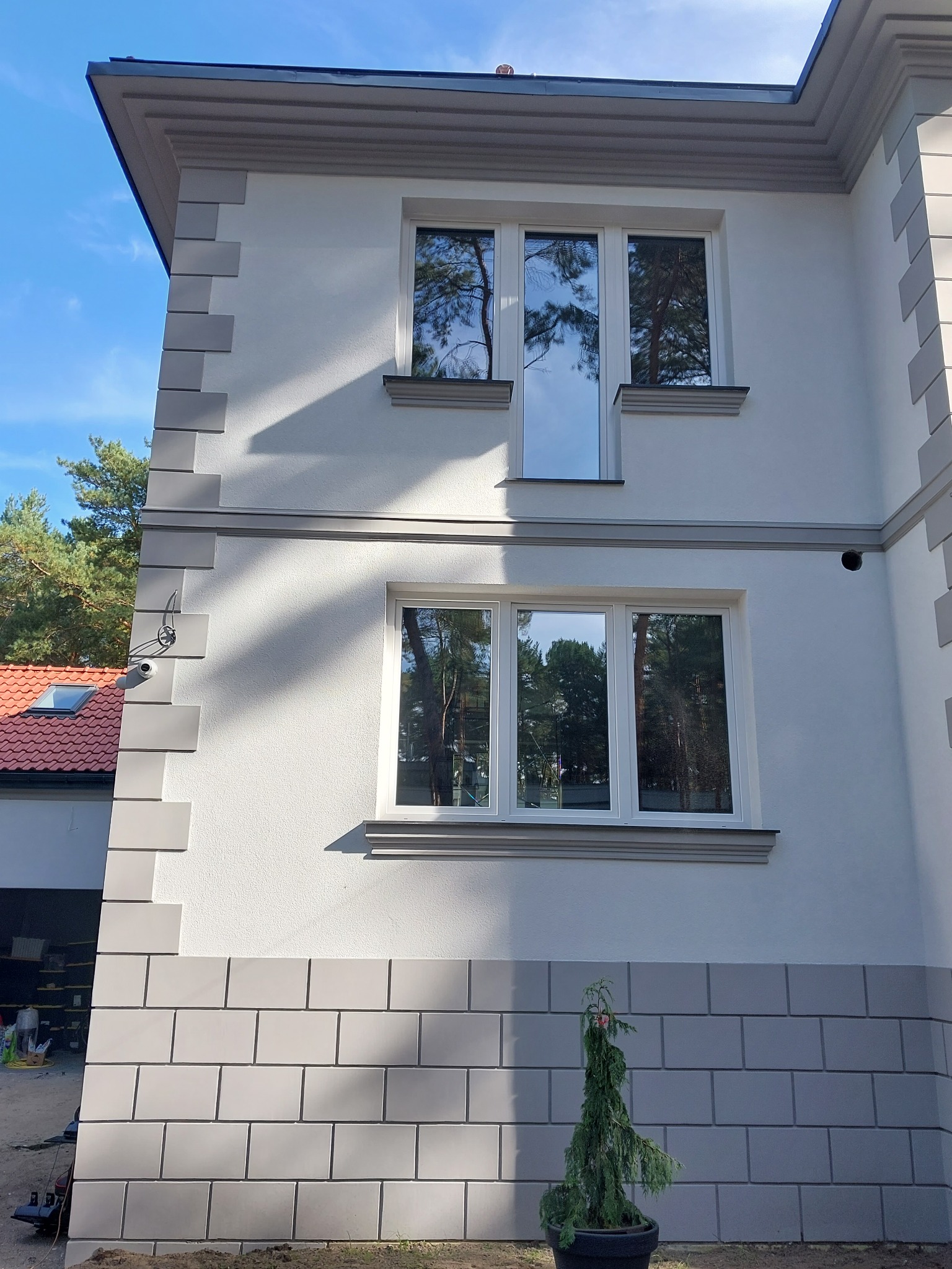 Elewacja domu z dekoracyjnym tynkiem imitującym cegłę, ozdobnymi gzymsami i oknami z odbijającym się lasem.