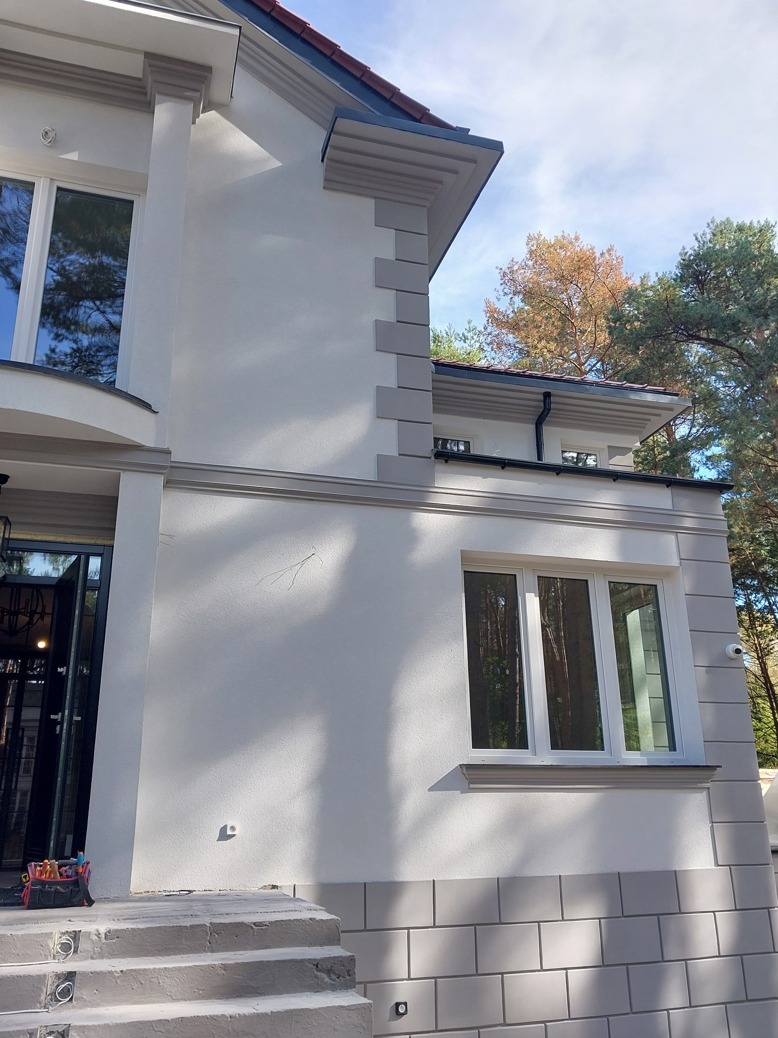 Elewacja dwukondygnacyjnego domu w Warszawie po tynkowaniu i malowaniu, z widocznymi detalami architektonicznymi i nowymi oknami, sfotografowana w słoneczny dzień.