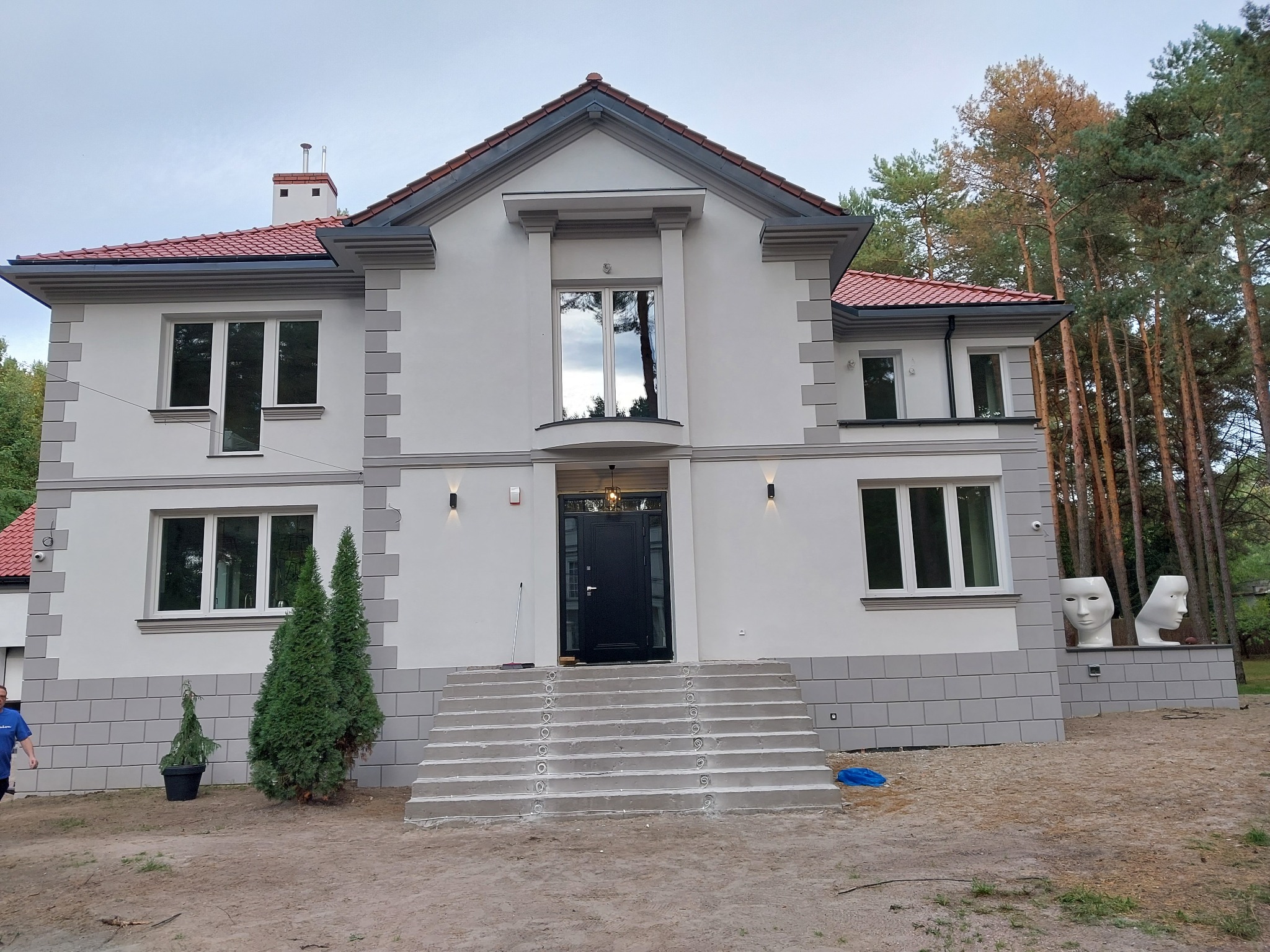 Elewacja dwukondygnacyjnego domu z szarymi detalami, czerwonym dachem i schodami wejściowymi, otoczona zielenią.