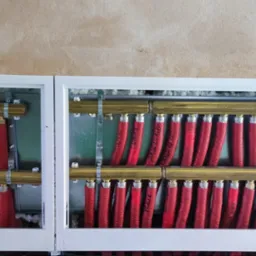 Rozdzielacz ogrzewania podłogowego w białej szafce, widoczne czerwone rury i mosiężne kolektory, surowa ściana w tle.