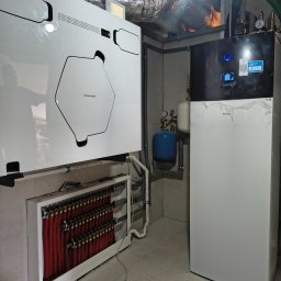 JMT-BUD Łukasz Pozorski - Nowoczesna instalacja grzewczo-wentylacyjna: biały rekuperator Thesslagreen, pompa ciepła Daikin, rozdzielacz ogrzewania podłogowego z czerwonymi rurami, srebrne kanały wentylacyjne.
