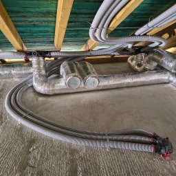 JMT-BUD Łukasz Pozorski - Szczegółowe ujęcie instalacji rekuperacji z elastycznymi rurami wentylacyjnymi i izolowanymi kanałami, biegnącej pod zielonymi deskami stropu i drewnianymi belkami, z częścią rur ułożonych...