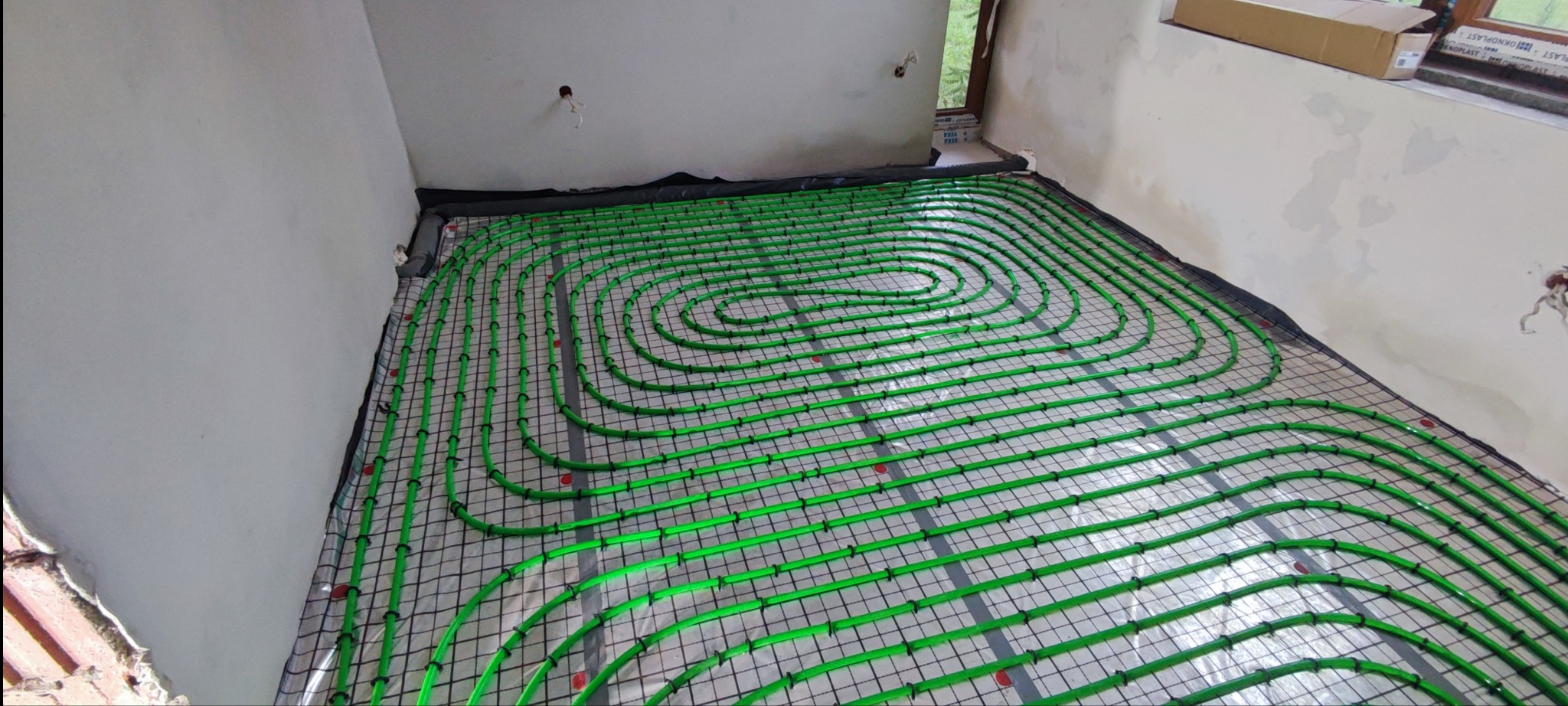 Zielone rurki ogrzewania podłogowego ułożone spiralnie na czarnej siatce w surowym wnętrzu przed wylewką.