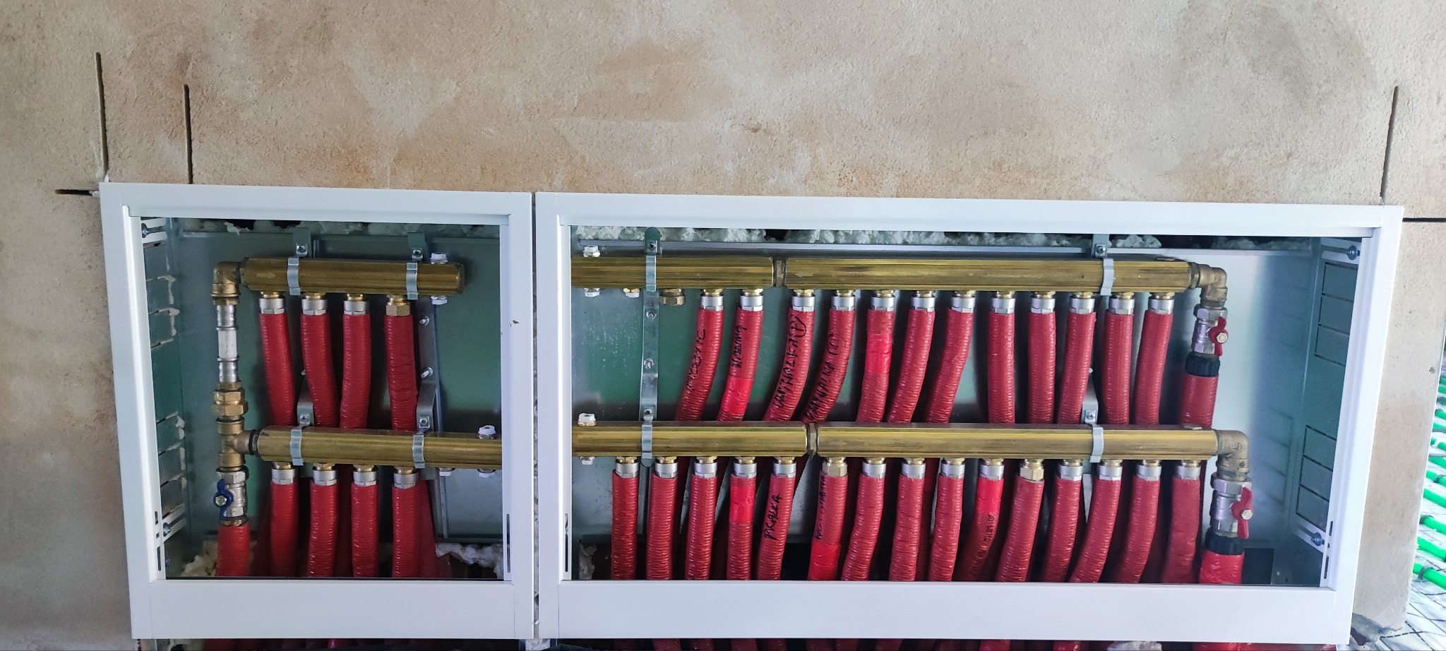 Rozdzielacz ogrzewania podłogowego w białej szafce, widoczne czerwone rury i mosiężne kolektory, surowa ściana w tle.