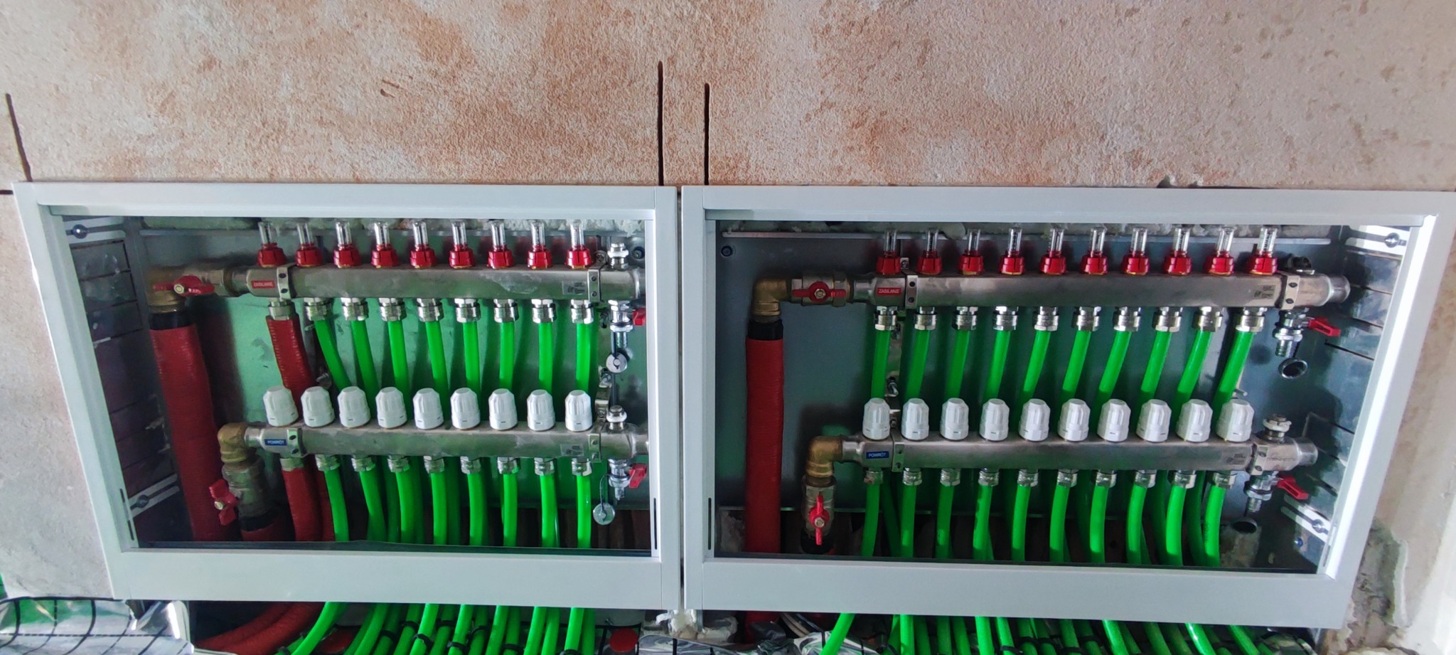 Rozdzielacz ogrzewania podłogowego w szafce, z zielonymi rurami, widoczne zawory i odpowietrzniki, zainstalowany na tle surowej ściany.