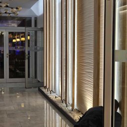 CICH-BUD CEZARY CICHOSZ - Montaż podświetlanej, dekoracyjnej ściany z drewnianymi panelami i falistą strukturą, z drobnymi białymi kamieniami u podstawy, widoczny pracownik w trakcie instalacji.