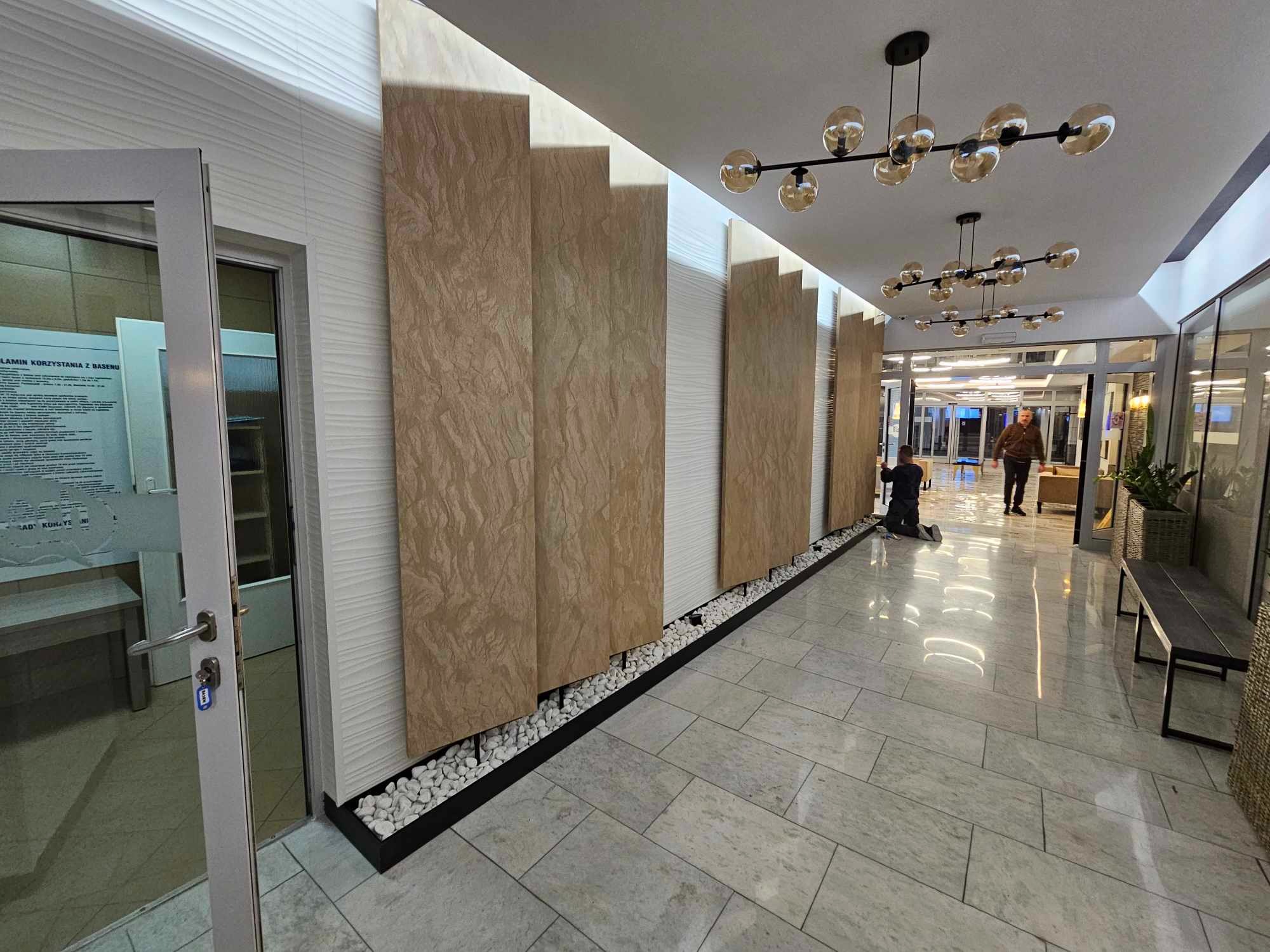 Montaż paneli dekoracyjnych w holu, widoczne pionowe płyty z teksturą kamienia, białe panele ścienne z delikatnym wzorem fal, dekoracyjne białe kamienie u podstawy ściany, eleganckie oświetlenie...