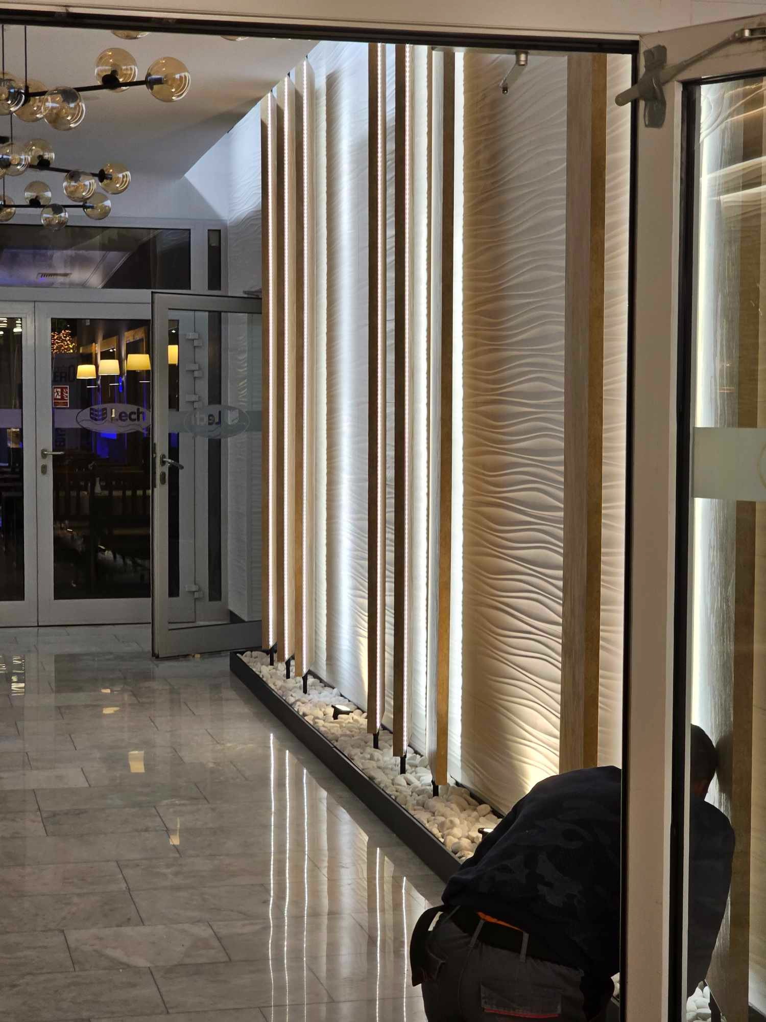 Montaż podświetlanej, dekoracyjnej ściany z drewnianymi panelami i falistą strukturą, z drobnymi białymi kamieniami u podstawy, widoczny pracownik w trakcie instalacji.