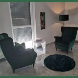 Wnętrze gabinetu z dwoma zielonymi fotelami typu uszak, okrągłym dywanem, stolikiem z chusteczkami higienicznymi i lampą podłogową, oświetlonego światłem dziennym wpadającym przez okno z żaluzjami.