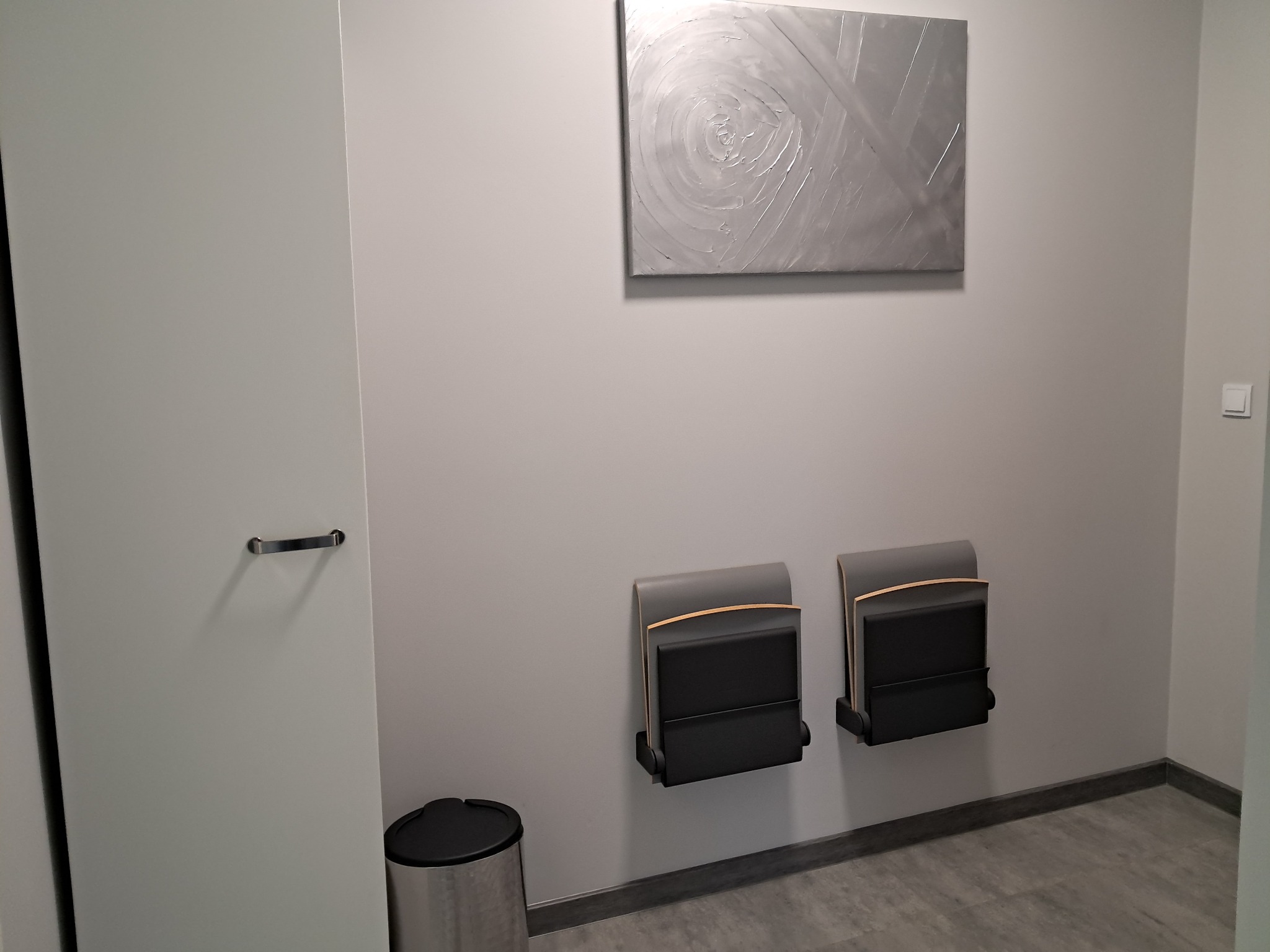 Poczekalnia w gabinecie z dwoma składanymi krzesłami ściennymi, srebrnym koszem na śmieci i abstrakcyjnym obrazem na ścianie.