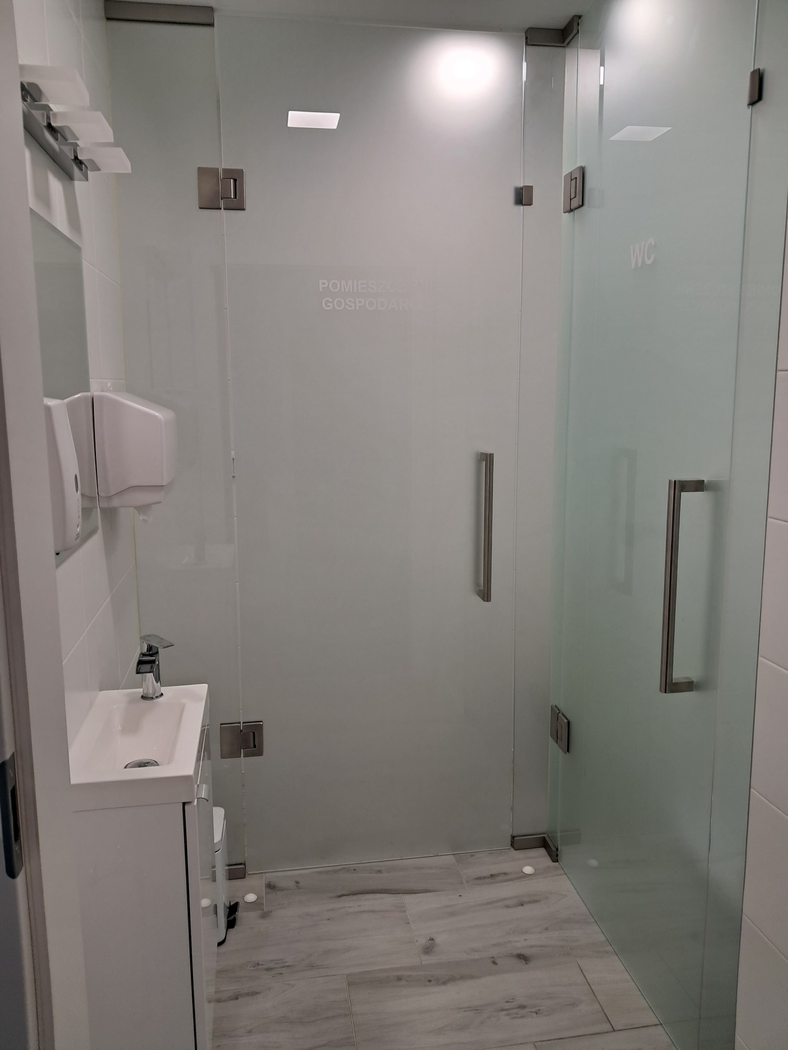 Nowoczesna toaleta z przeszklonymi drzwiami i minimalistyczną umywalką z dozownikiem mydła, widoczne napisy 'Pomieszczenie Gospodarcze' i 'WC' na szkle.