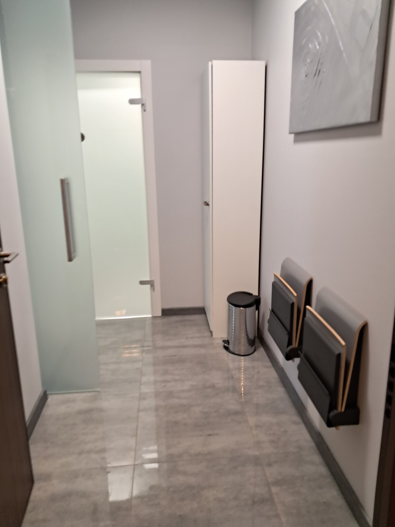 Wnętrze gabinetu z szarą podłogą, białymi ścianami, minimalistycznym obrazem, szafką, koszem na śmieci i składanymi siedziskami przy ścianie.