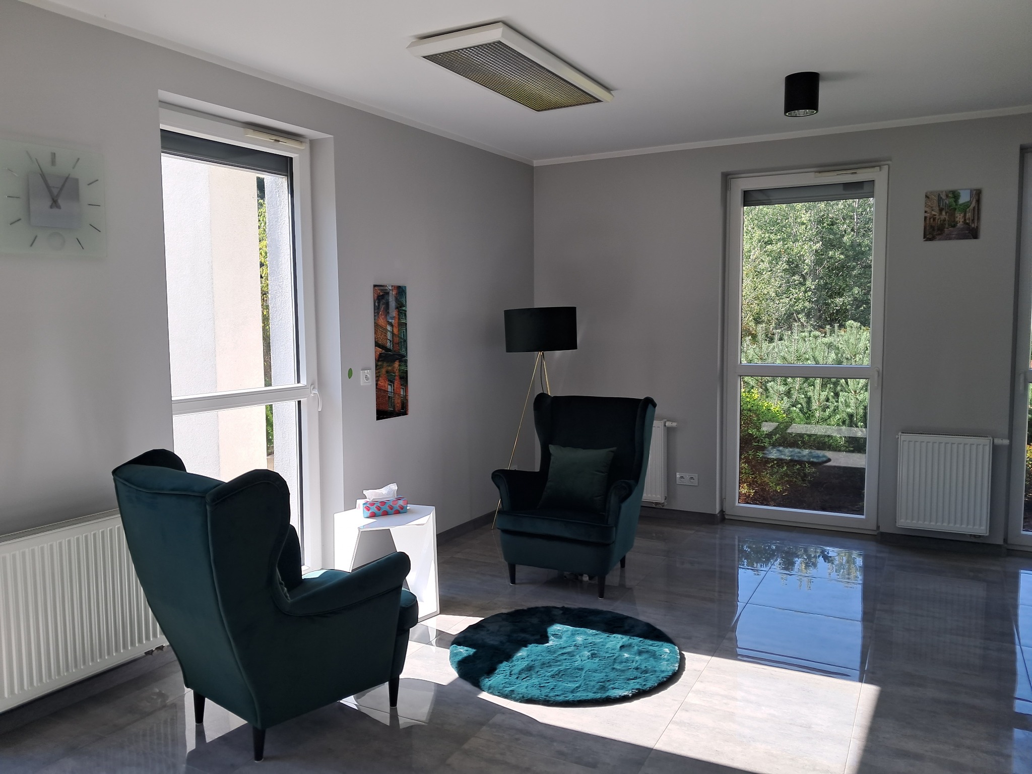 Jasne wnętrze gabinetu z dwoma zielonymi fotelami typu uszak, okrągłym turkusowym dywanikiem, białym stolikiem, zegarem na ścianie i dużymi oknami wpuszczającymi naturalne światło.