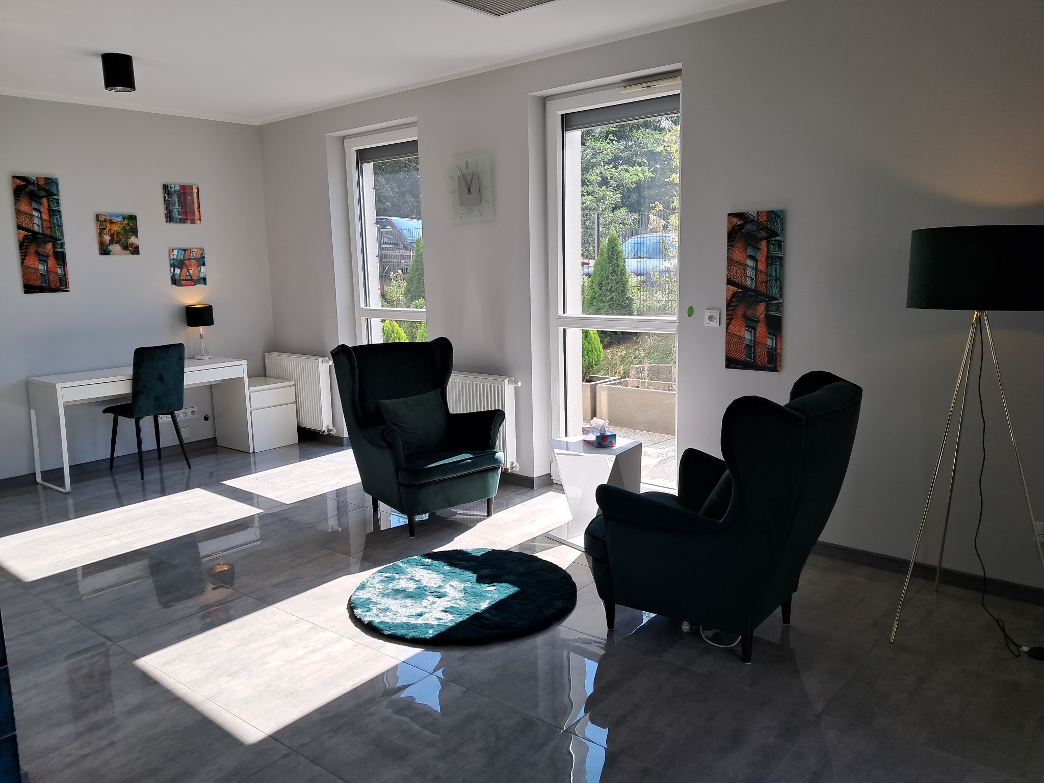 Jasne wnętrze gabinetu z dwoma zielonymi fotelami typu uszak, okrągłym dywanem, biurkiem i lampą na trójnogu, udekorowane obrazami z motywami miejskimi na ścianach.