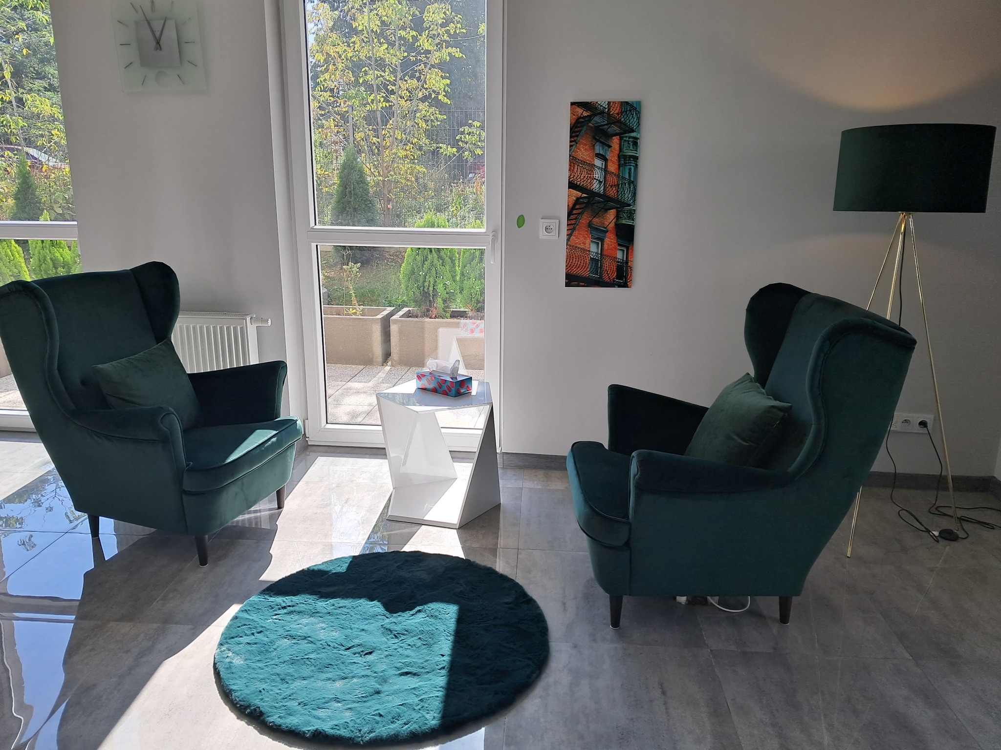 Jasny gabinet z dwoma zielonymi fotelami uszakami, okrągłym turkusowym dywanikiem, białym stolikiem i lampą na złotym statywie, obraz na ścianie przedstawia budynek z metalowymi schodami pożarowymi.