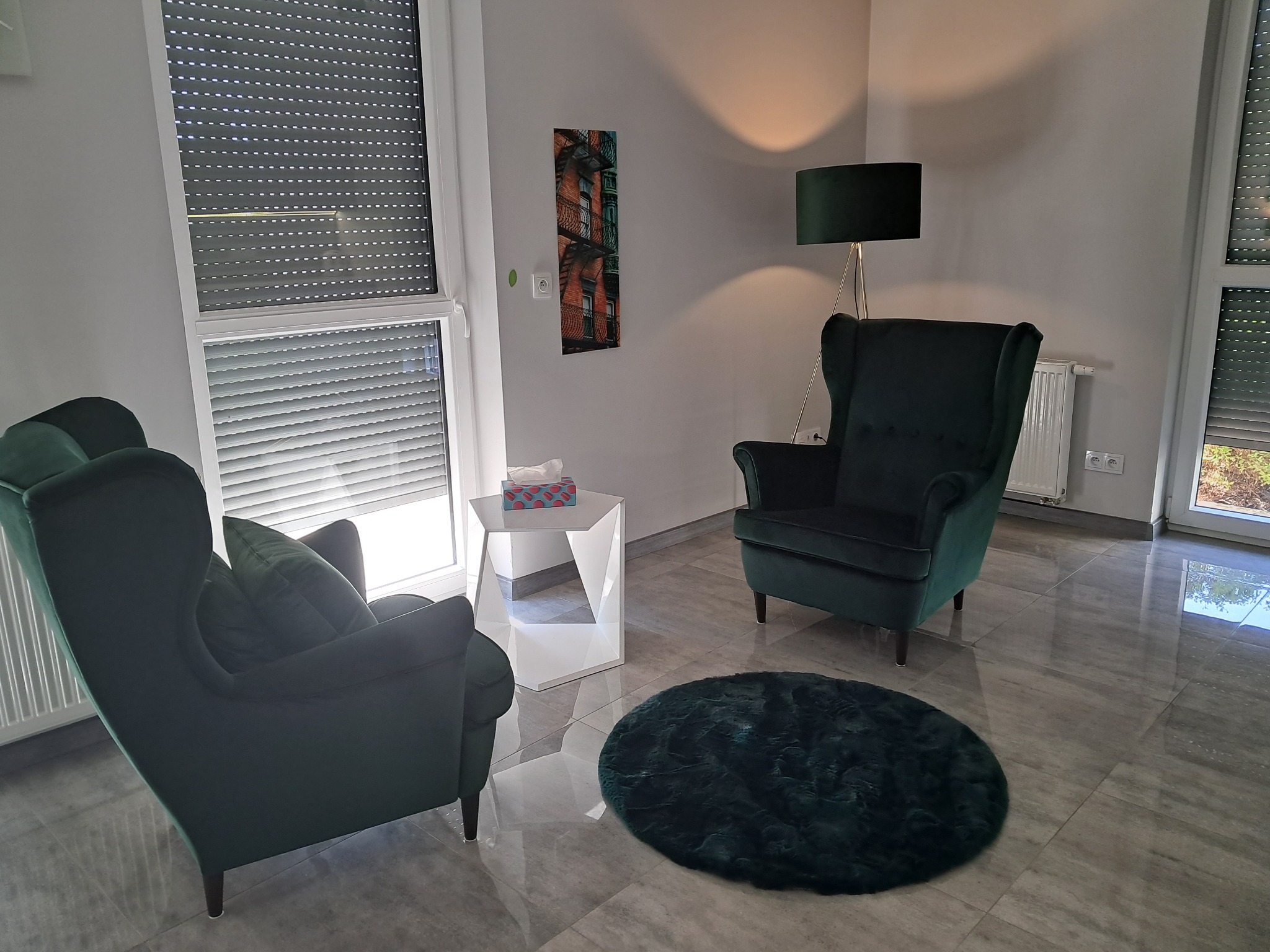 Gabinet z dwoma zielonymi fotelami typu uszak, okrągłym dywanem, lampą stojącą i stolikiem z chusteczkami, przytulne i stonowane wnętrze.