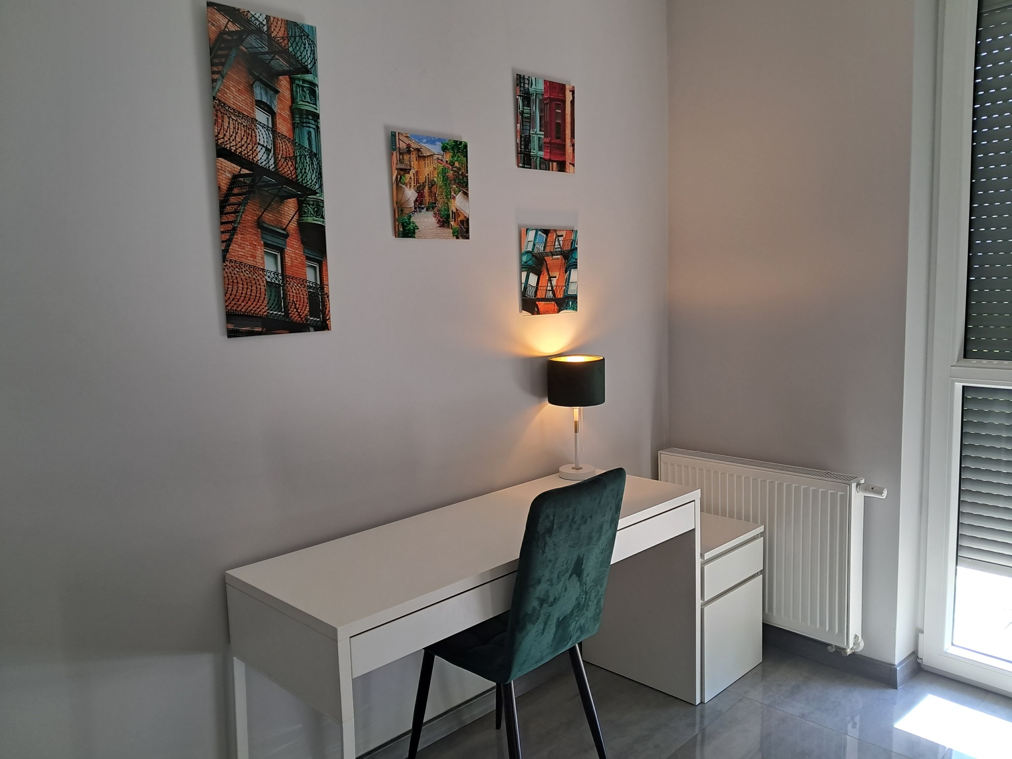 Nowoczesne biuro z białym biurkiem, zielonym krzesłem i lampką, ozdobione obrazami z motywami miejskimi na szarej ścianie.
