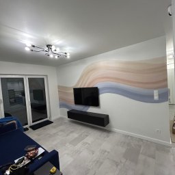 Miko-Art Usługi Remotowo-Wykończeniowe - Salon po odświeżeniu: ściana z abstrakcyjnym malunkiem w odcieniach beżu i błękitu, podwieszana szafka RTV i telewizor. Nowoczesne oświetlenie sufitowe.