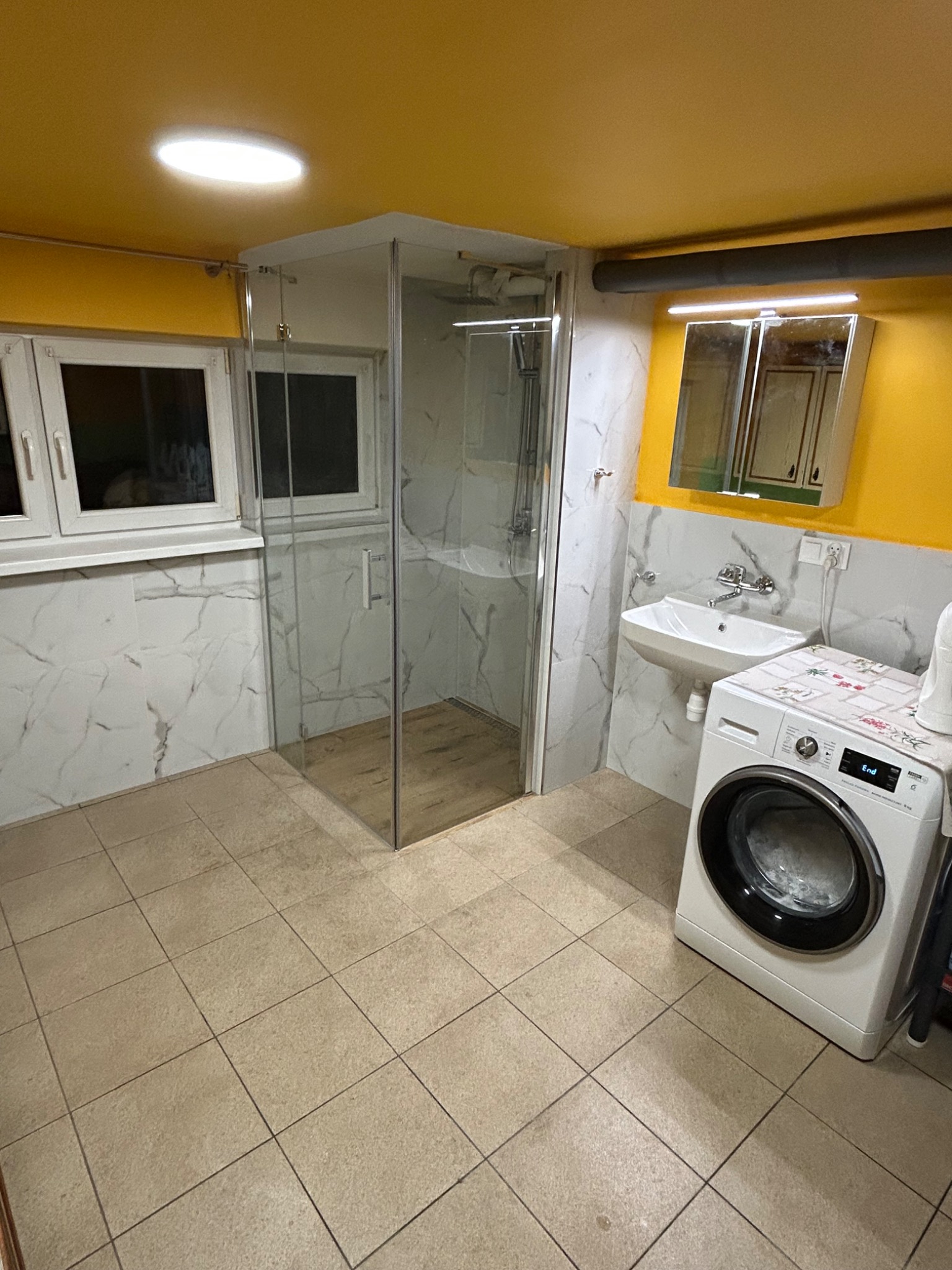 Wykończona łazienka z kabiną prysznicową, umywalką i pralką. Żółty sufit, białe marmurowe płytki ścienne i beżowa podłoga z płytek. Widoczne okno i lustro.