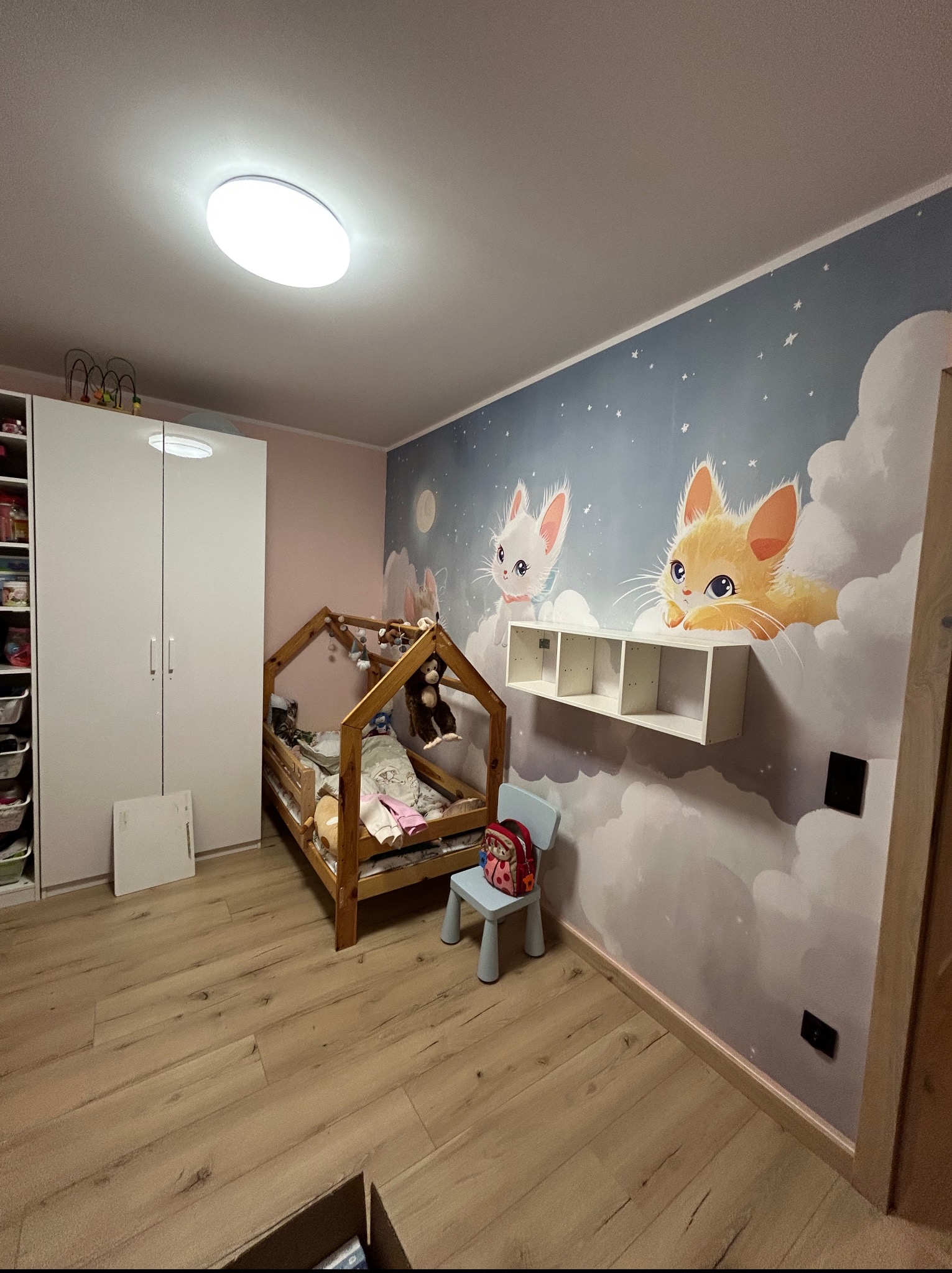 Pokój dziecięcy z łóżkiem w kształcie domku, szafą i ścianą z malowidłami kotów na tle chmur. Podłoga wyłożona panelami imitującymi drewno.