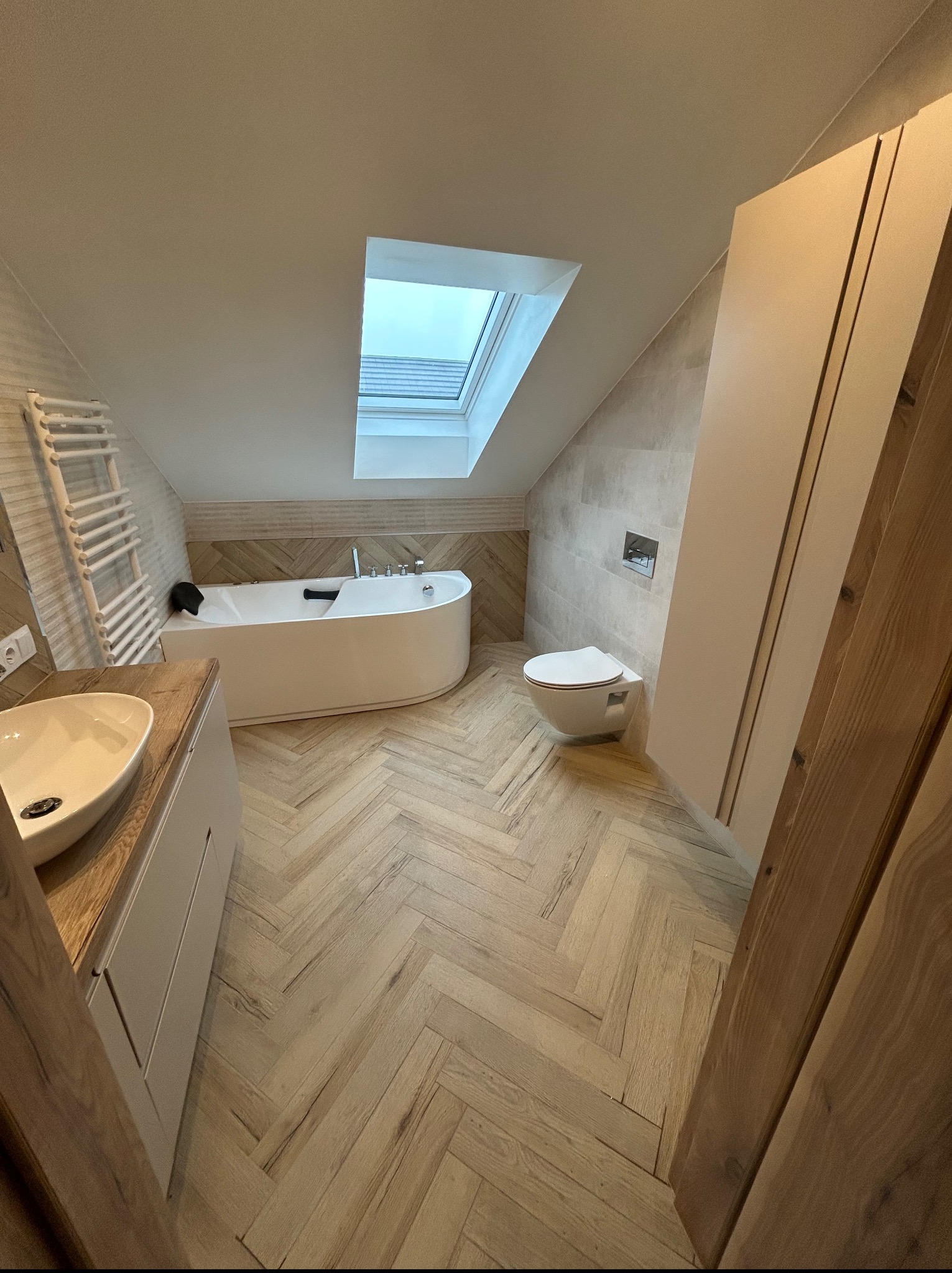 Jasna łazienka na poddaszu z oknem dachowym, wykończona w beżach i bieli. Umywalka z szafką, wanna, toaleta i grzejnik drabinkowy. Parkiet ułożony w jodełkę.