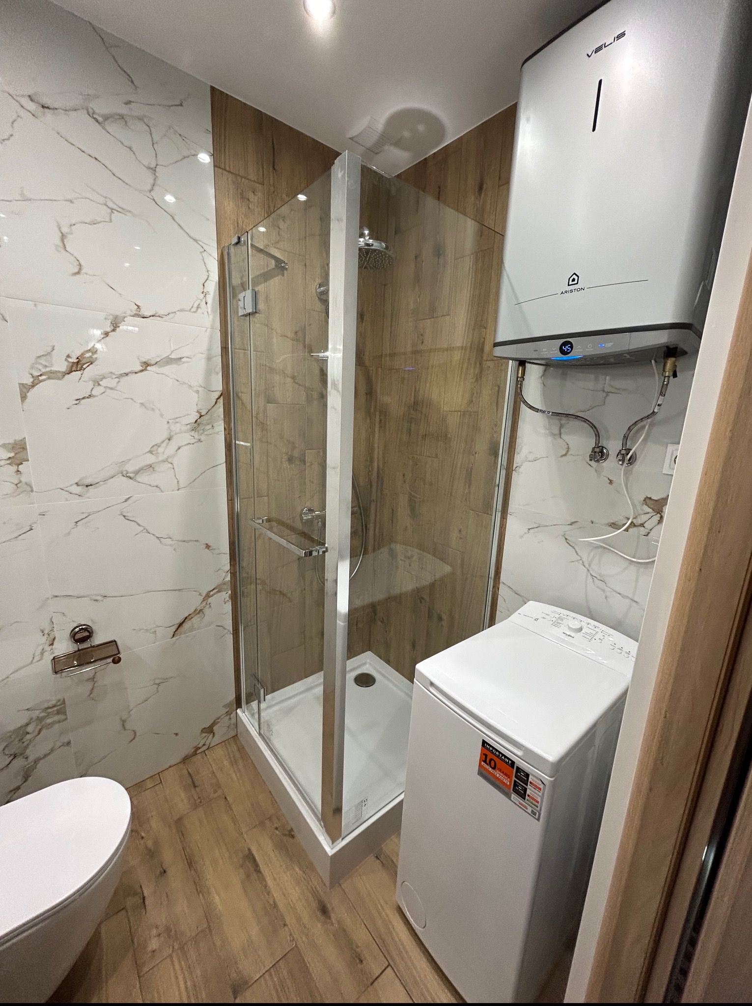 Nowoczesna łazienka po remoncie: kabina prysznicowa, marmurowe i drewnopodobne płytki, biała pralka i bojler Ariston Velis. Widok na całe pomieszczenie.
