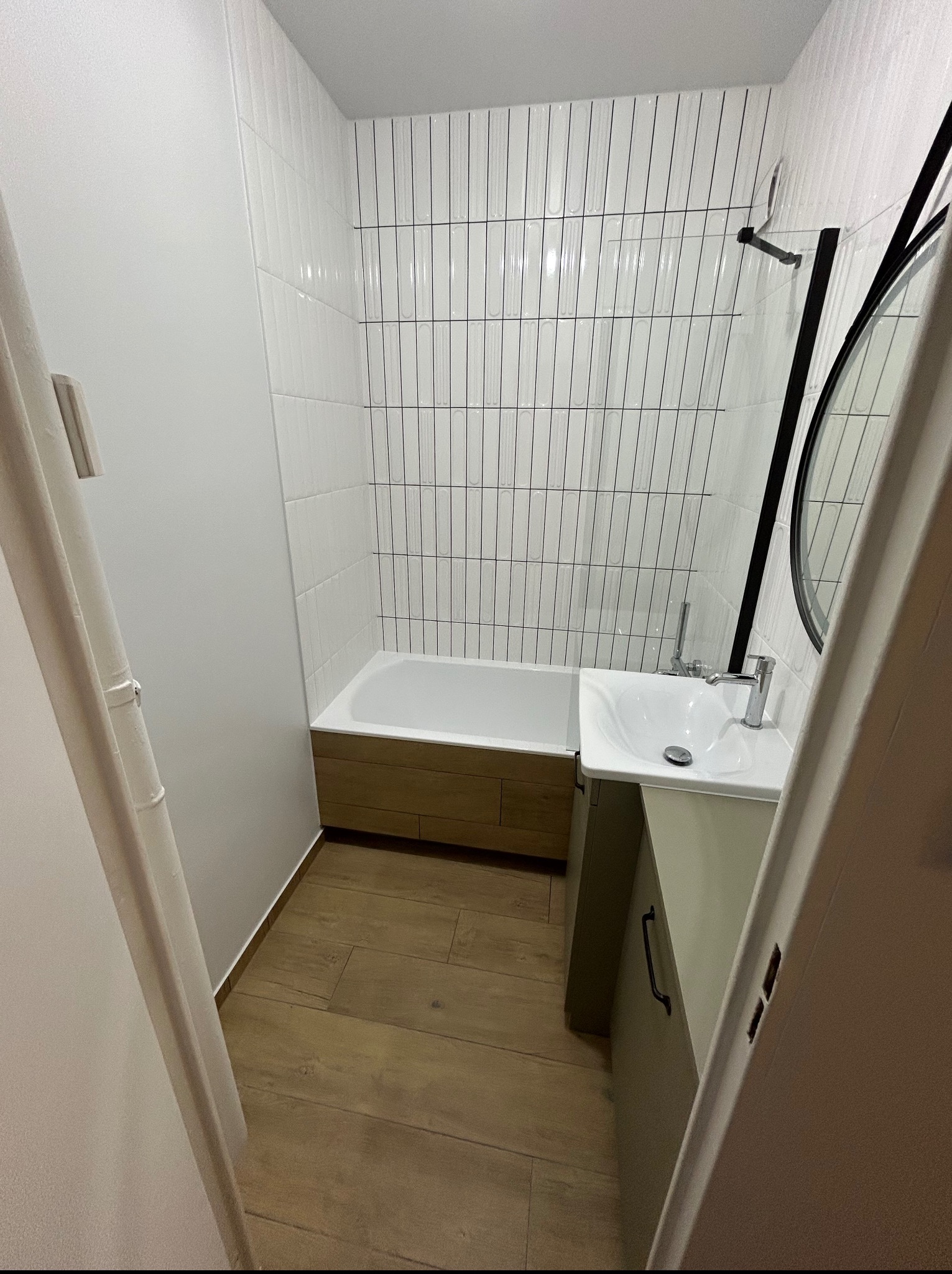 Nowoczesna łazienka po remoncie: białe, pionowe płytki na ścianie, drewnopodobna podłoga, wanna z obudową i umywalka z szafką. Minimalistyczny design.