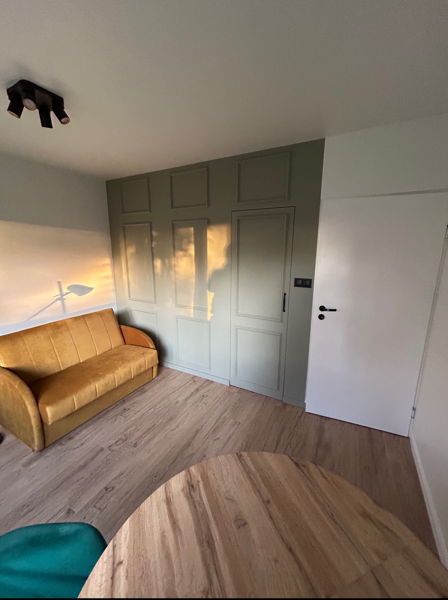 Pokój z panelami podłogowymi i ścianą w odcieniach szarości z dekoracyjnymi panelami. W tle sofa w kolorze musztardowym i cień rzucany przez lampę.