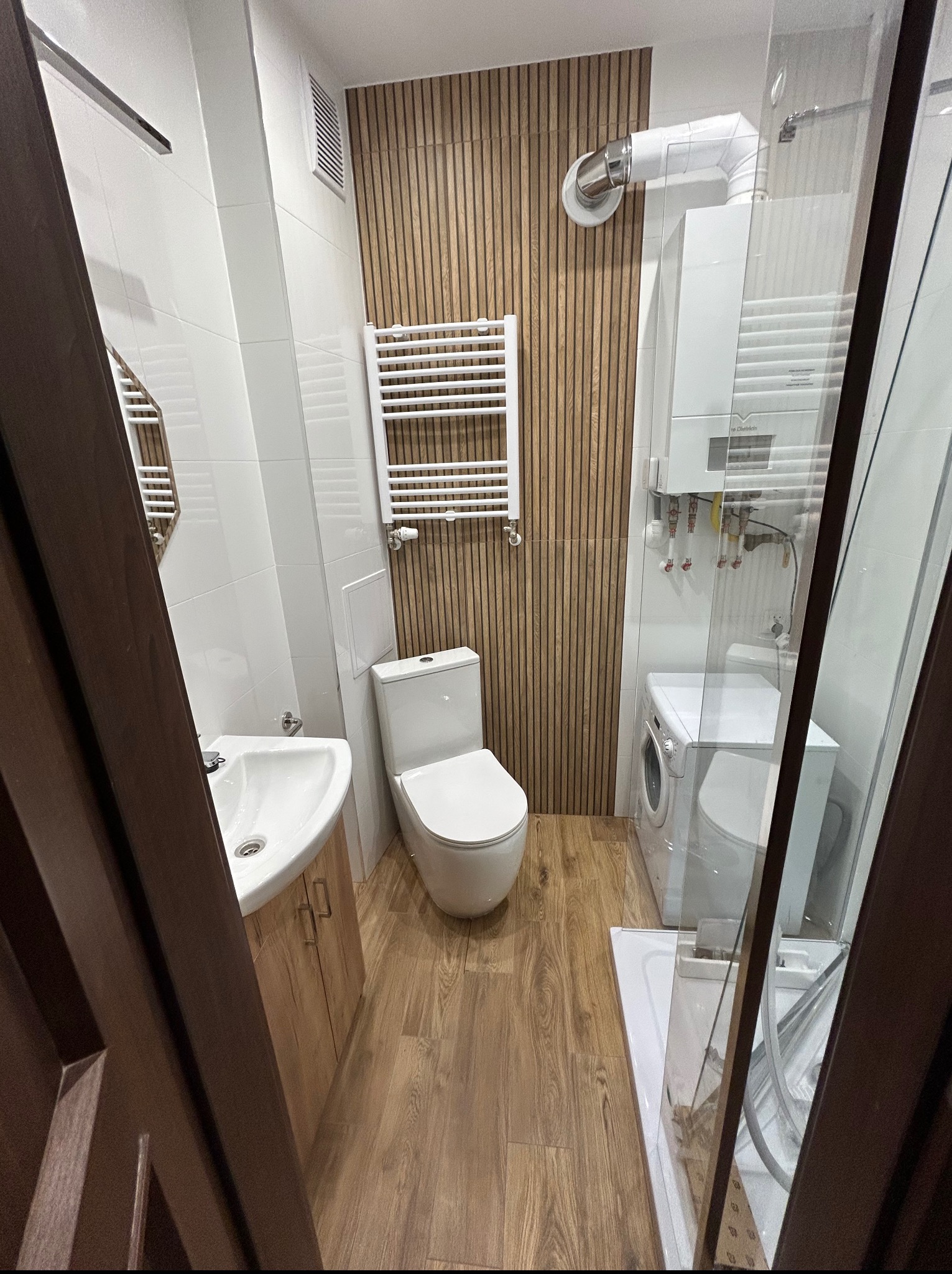 Nowoczesna łazienka z drewnianymi panelami ściennymi, białymi płytkami, umywalką z szafką, toaletą i kabiną prysznicową. Widoczny bojler i pralka. Całość utrzymana w jasnej kolorystyce.