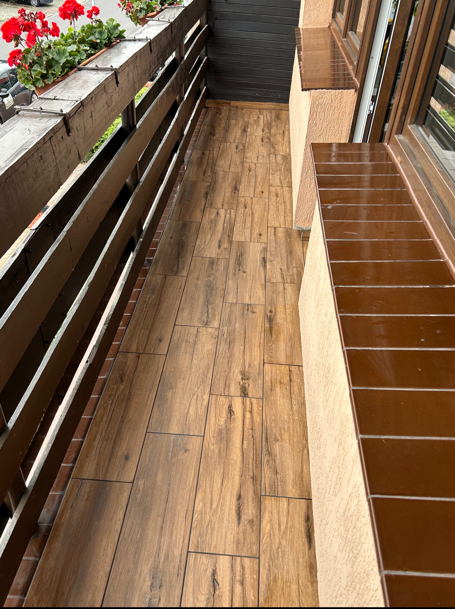 Wykończony balkon z drewnopodobnymi płytkami na podłodze, brązową balustradą i donicami z czerwonymi kwiatami. Widok z góry, ukazujący detale wykończenia.