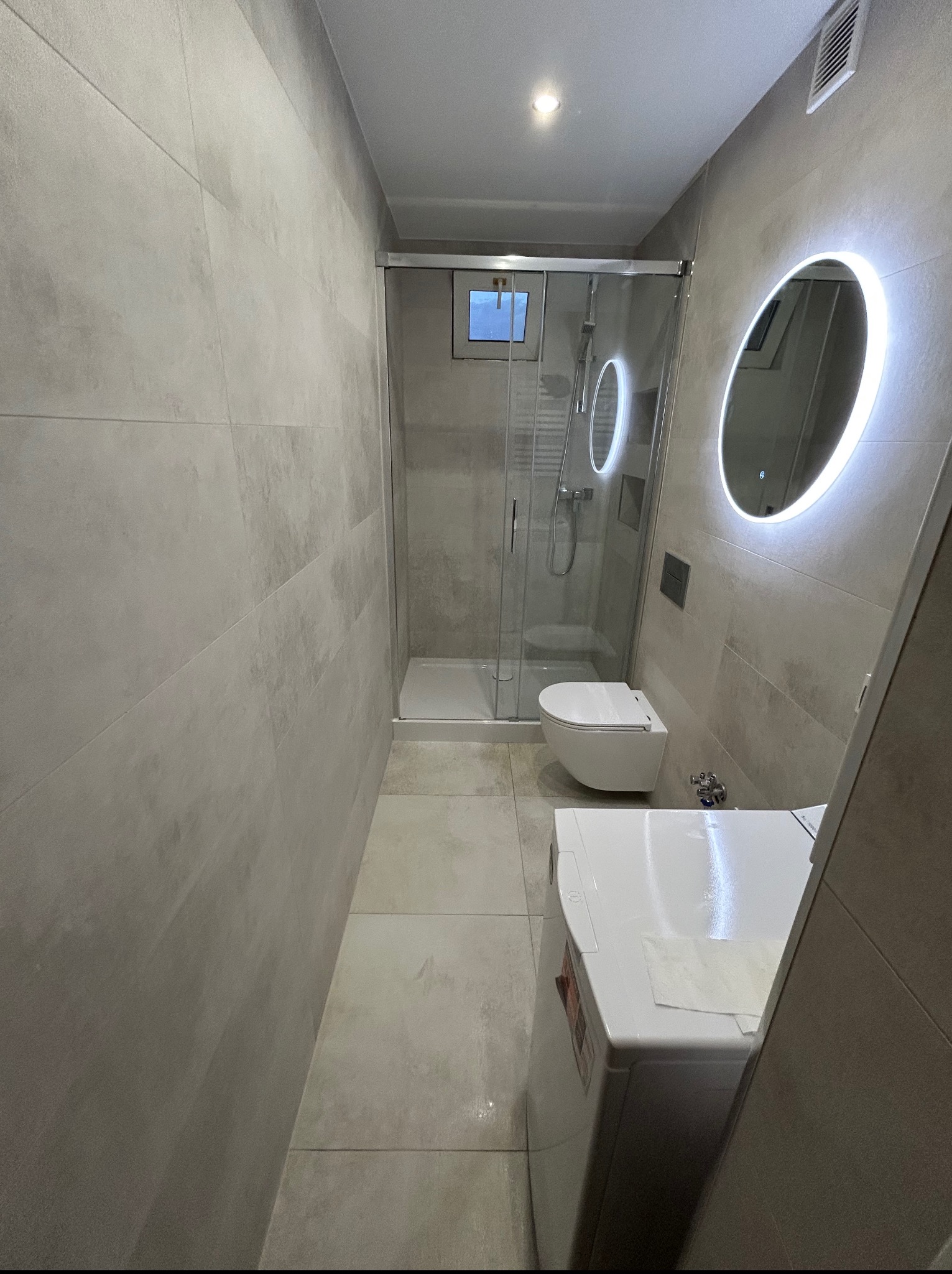 Nowoczesna łazienka z kabiną prysznicową, podświetlanym lustrem i pralką. Szare płytki na ścianach i podłodze. Minimalistyczny design, czysta przestrzeń.