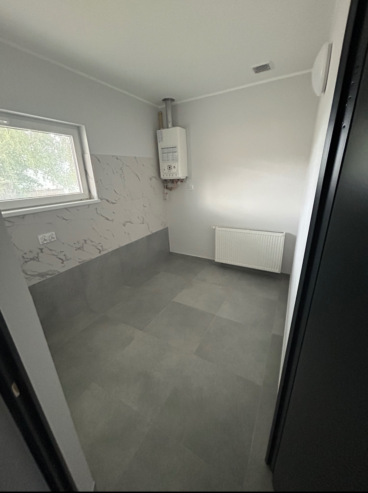 Wykończona łazienka w szarościach: płytki podłogowe i ścienne imitujące marmur, biały grzejnik, piec gazowy. Widok zza czarnej ramy drzwi.