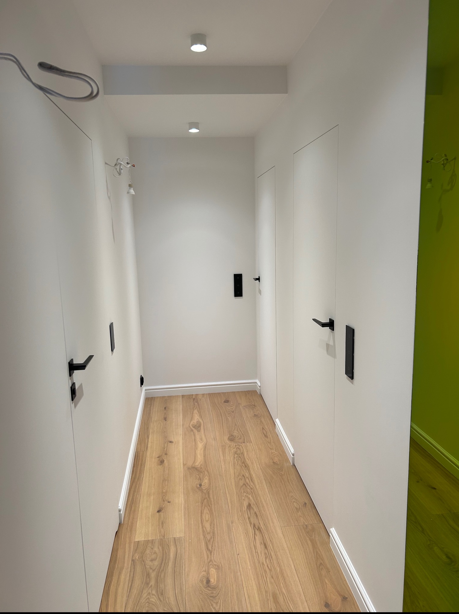 Biały korytarz z drewnianą podłogą, czarne klamki i minimalistyczne oświetlenie punktowe. Ściany gładkie, z ukrytymi drzwiami i zielonym akcentem kolorystycznym w tle.