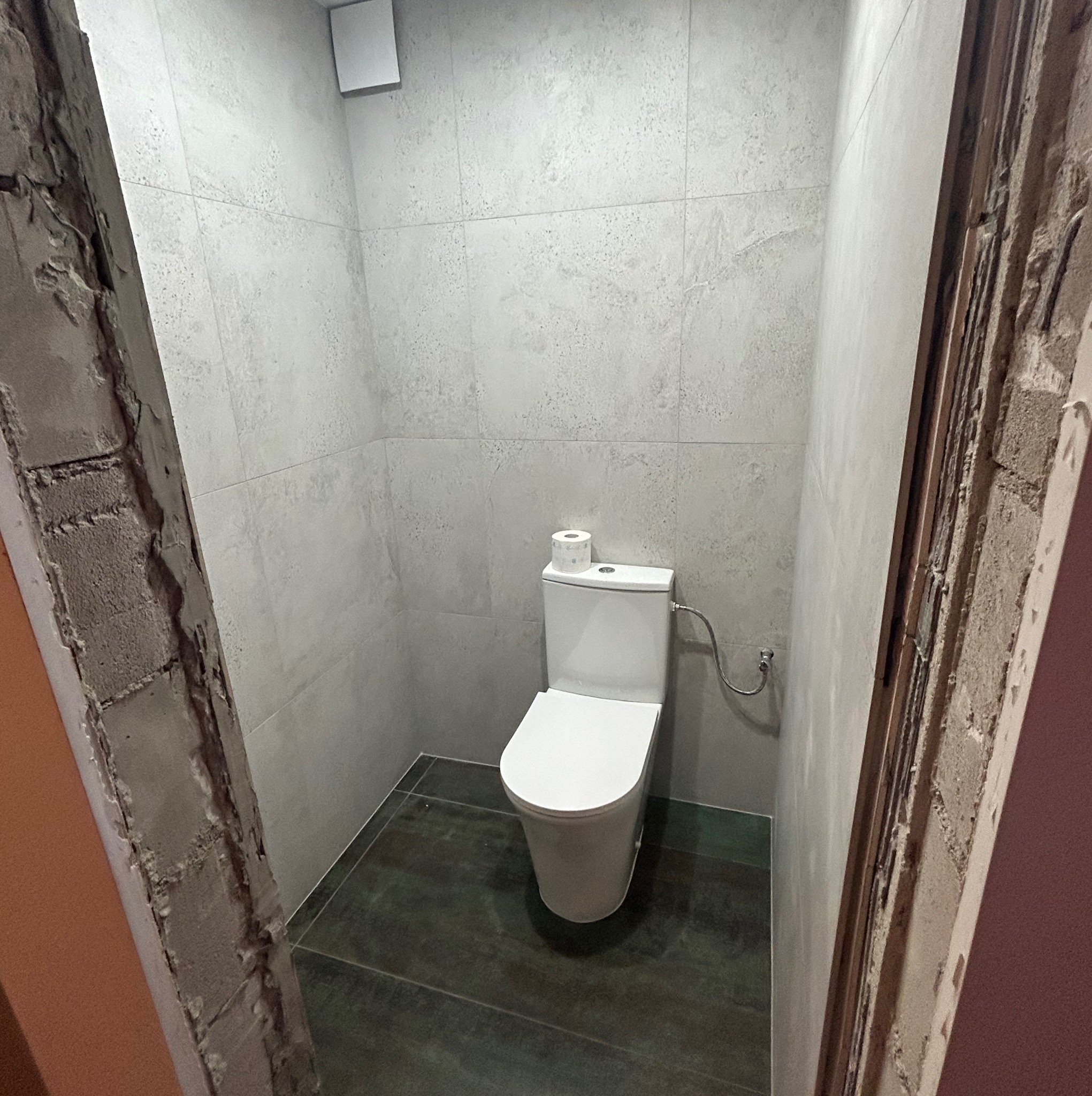 Wykończona łazienka z toaletą, szarymi płytkami na ścianach i podłodze. Widoczne surowe wykończenie ościeżnicy drzwi, kontrastujące z nowoczesnym wnętrzem.