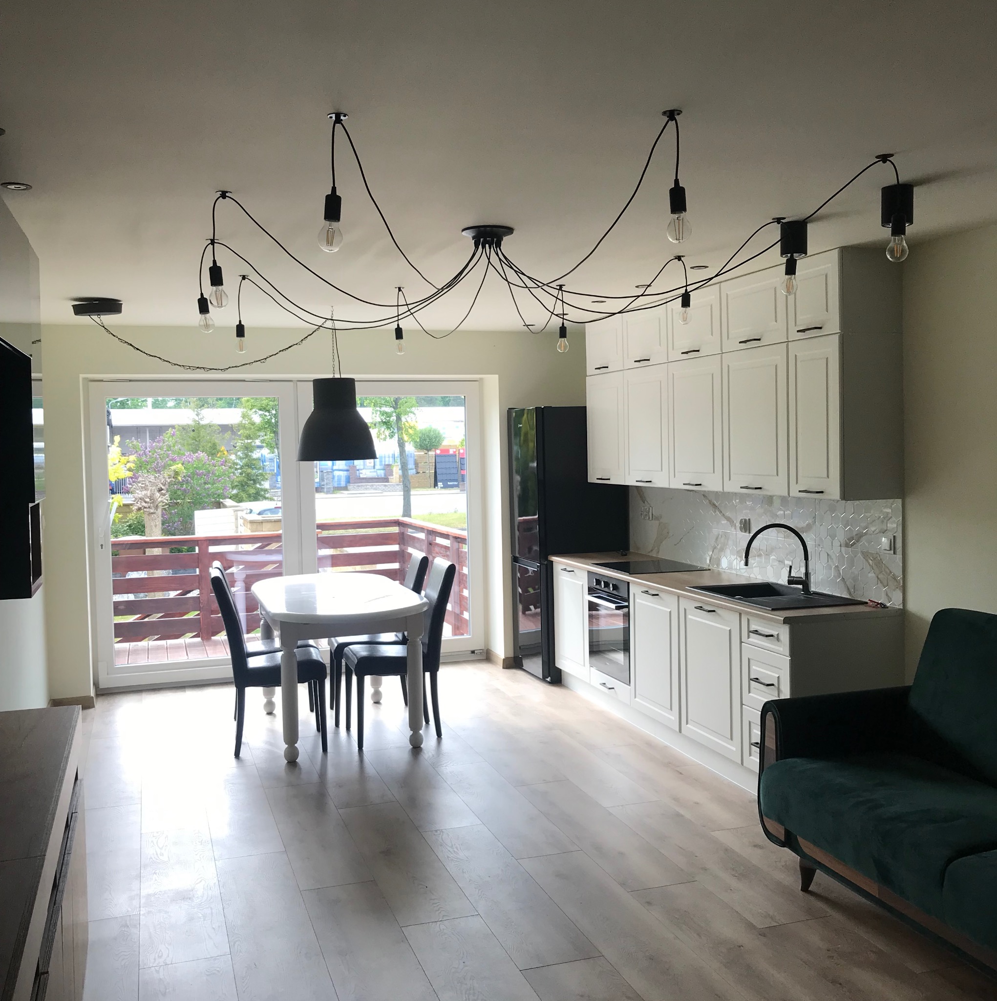 Jasne wnętrze z białą kuchnią, czarnymi akcentami i oryginalnym oświetleniem. Widoczne panele podłogowe, stół z krzesłami i fragment zielonej sofy.