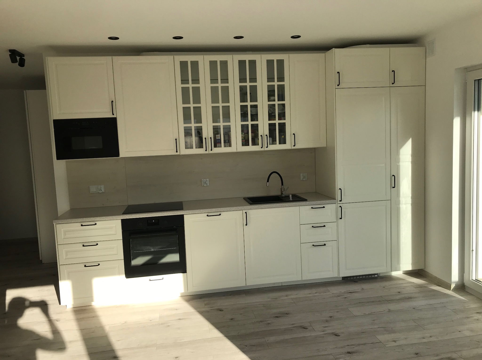 Biała zabudowa kuchenna z czarnymi uchwytami i sprzętami AGD, widok na szafki z przeszkleniami i drewnopodobną podłogę w nowym wnętrzu.