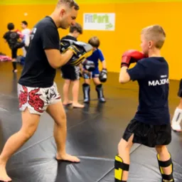 Instruktor z czarną koszulką i wzorzystych spodenkach trzyma tarcze treningowe, podczas gdy młody chłopak w ochraniaczach i rękawicach bokserskich ćwiczy ciosy na tle żółtej ściany w sali treningowej.