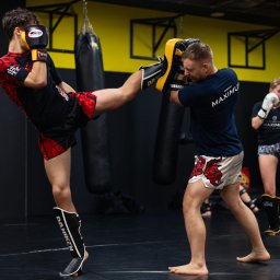 Mariusz Kudłak - Mężczyzna w czarno-czerwonym stroju kopie w tarczę trzymaną przez trenera w ciemnej sali treningowej; w tle wiszące worki bokserskie i trenująca kobieta.