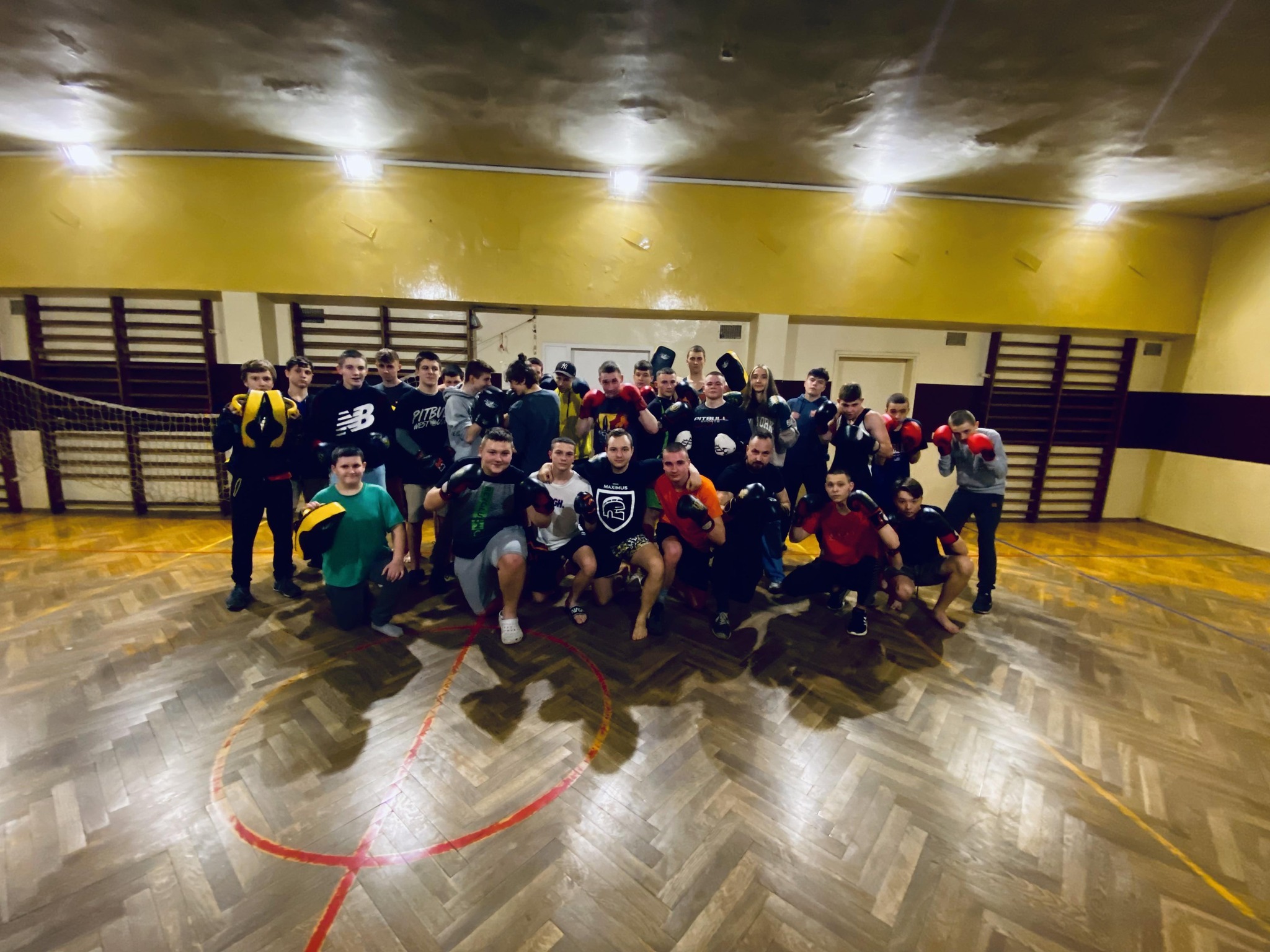 Grupa osób w strojach sportowych i rękawicach bokserskich pozuje do zdjęcia w sali gimnastycznej z parkietem w jodełkę i żółtymi ścianami.