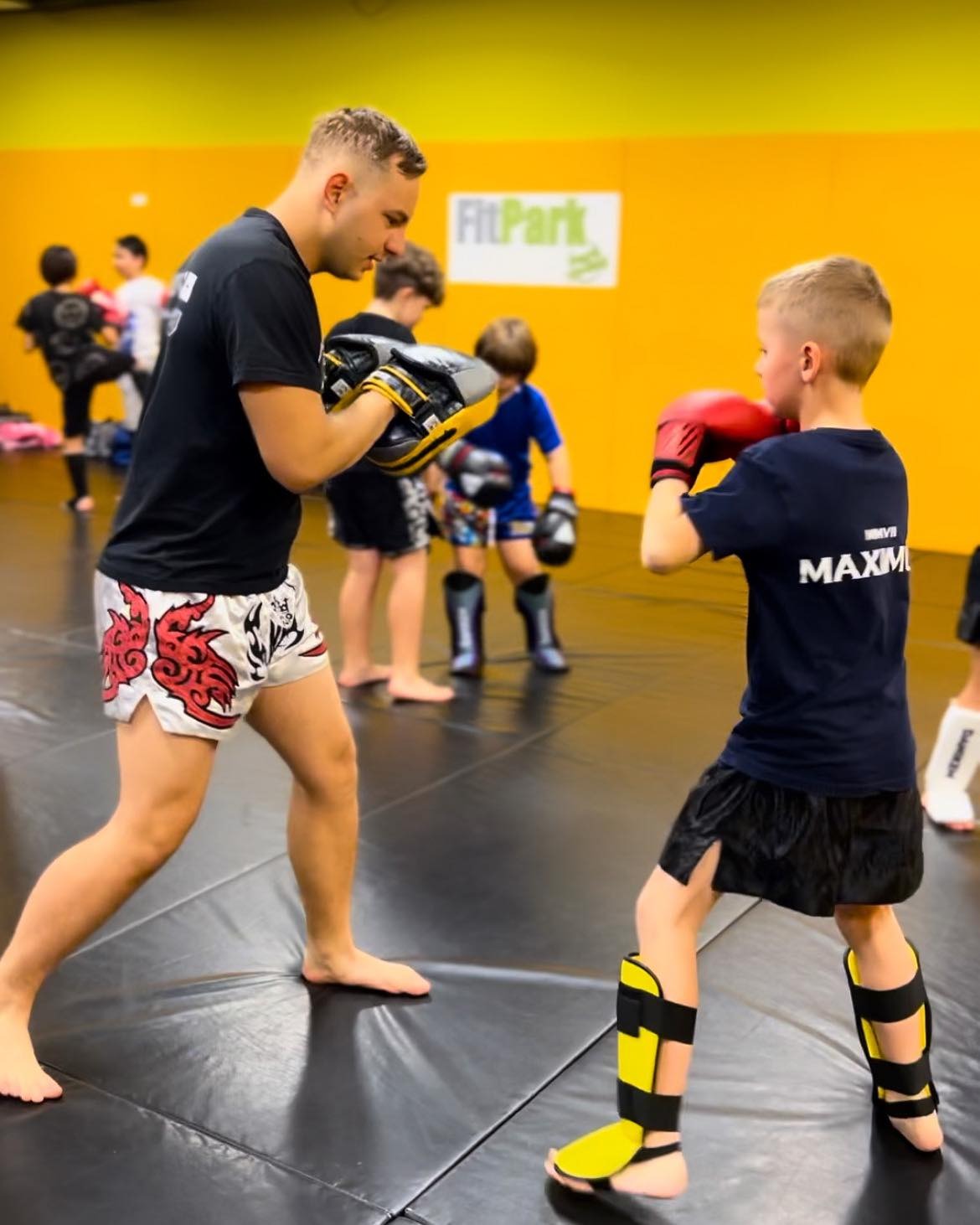 Instruktor z czarną koszulką i wzorzystych spodenkach trzyma tarcze treningowe, podczas gdy młody chłopak w ochraniaczach i rękawicach bokserskich ćwiczy ciosy na tle żółtej ściany w sali treningowej.