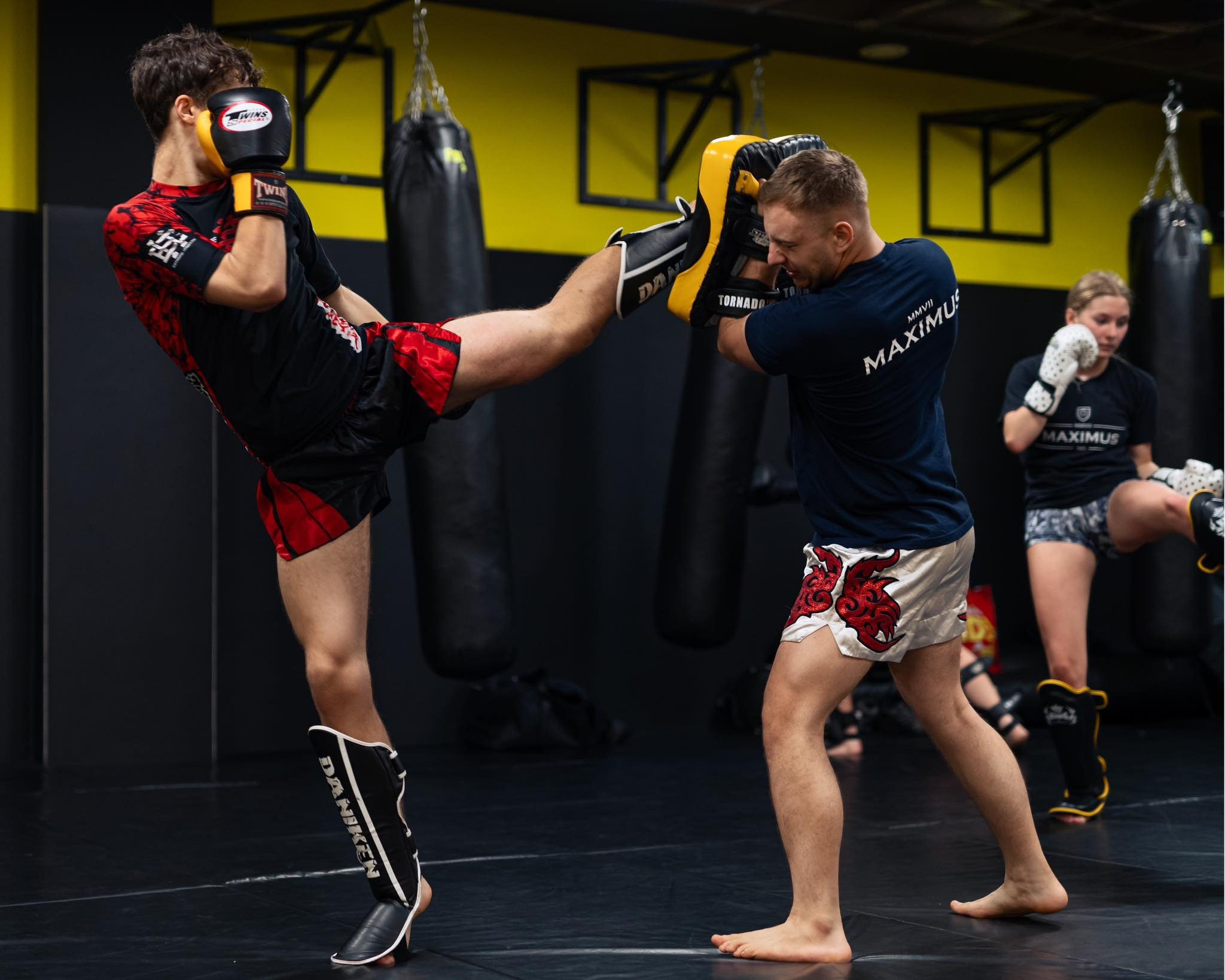 Mężczyzna w czarno-czerwonym stroju kopie w tarczę trzymaną przez trenera w ciemnej sali treningowej; w tle wiszące worki bokserskie i trenująca kobieta.