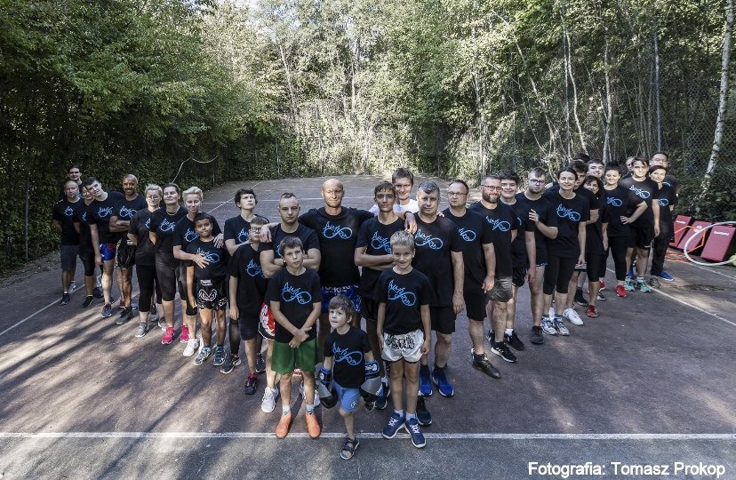 Grupa dorosłych i dzieci w czarnych koszulkach z logo stoi na zewnątrz na tle drzew, pozując do zdjęcia po treningu sportów walki.
