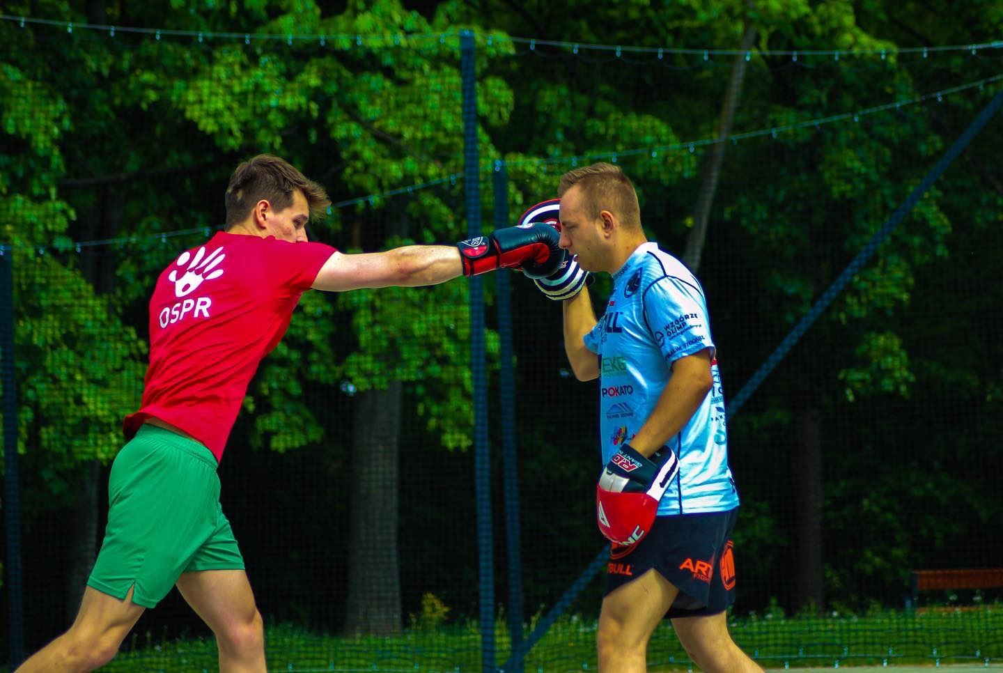 Dwóch mężczyzn w strojach sportowych i rękawicach bokserskich podczas sparingu na świeżym powietrzu, na tle zieleni.
