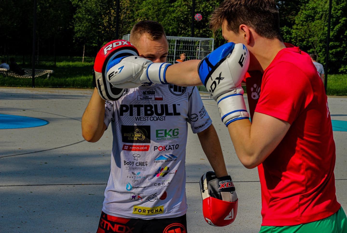 Trening sportów walki na zewnątrz: mężczyzna w białej koszulce z logotypami różnych firm uderza w tarczę trzymaną przez drugiego mężczyznę w czerwonej koszulce, obaj w rękawicach bokserskich RDX.